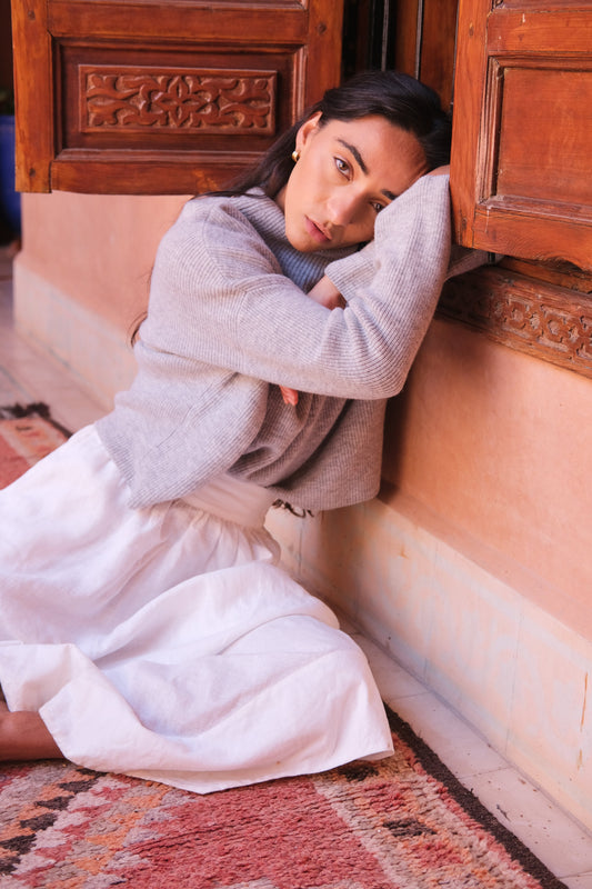 LILLY PILLY woman wearing a grey Lumi cashmere knit sweater leaning against a wooden door in a warm-toned interior setting.