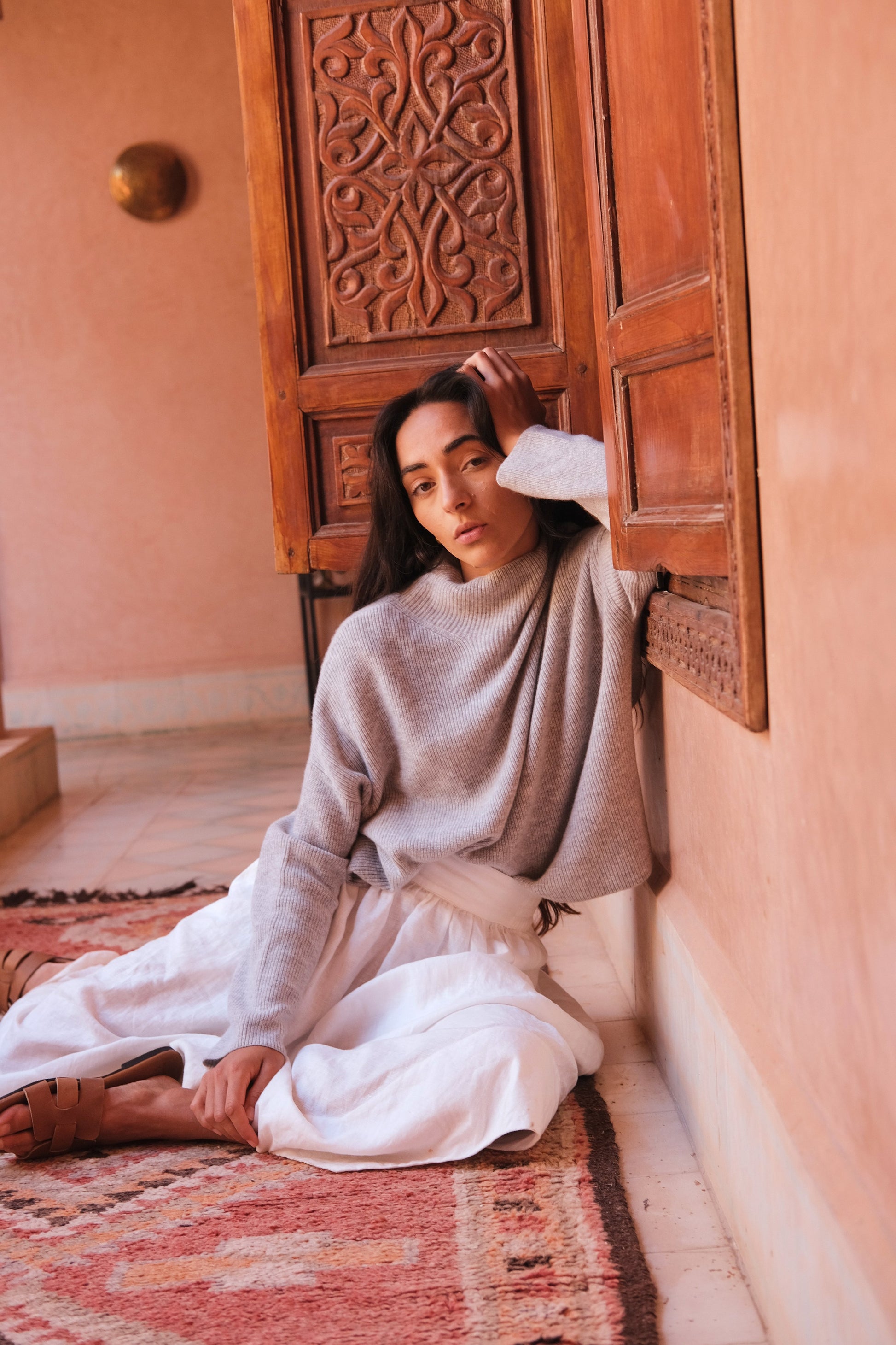 LILLY PILLY woman wearing a grey Lumi cashmere knit sweater and ivory white skirt  sitting on a patterned rug in a room with ornate wooden doors.