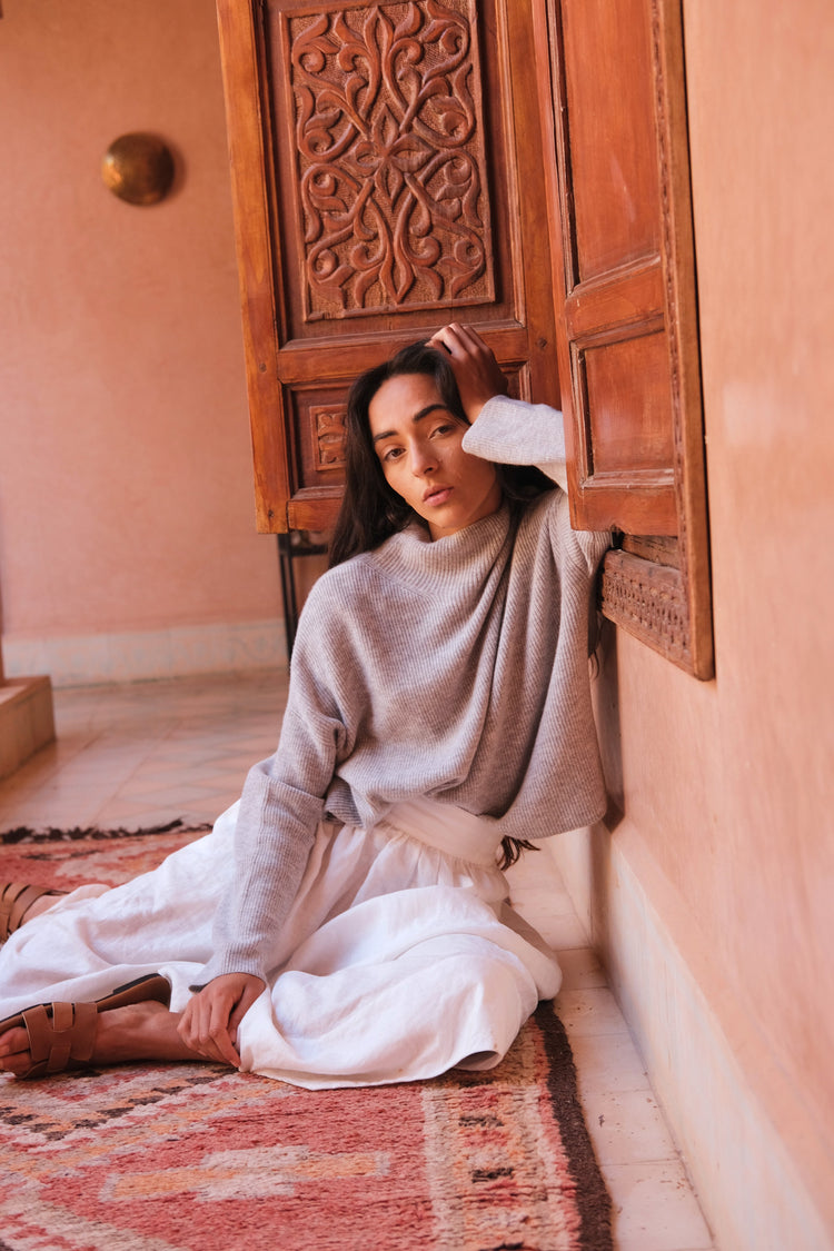 Lumi Cashmere Knit in Grey Marle - front view, styled with an ivory skirt, seated on a patterned rug in a room with ornate wooden doors - Lilly Pilly Collection