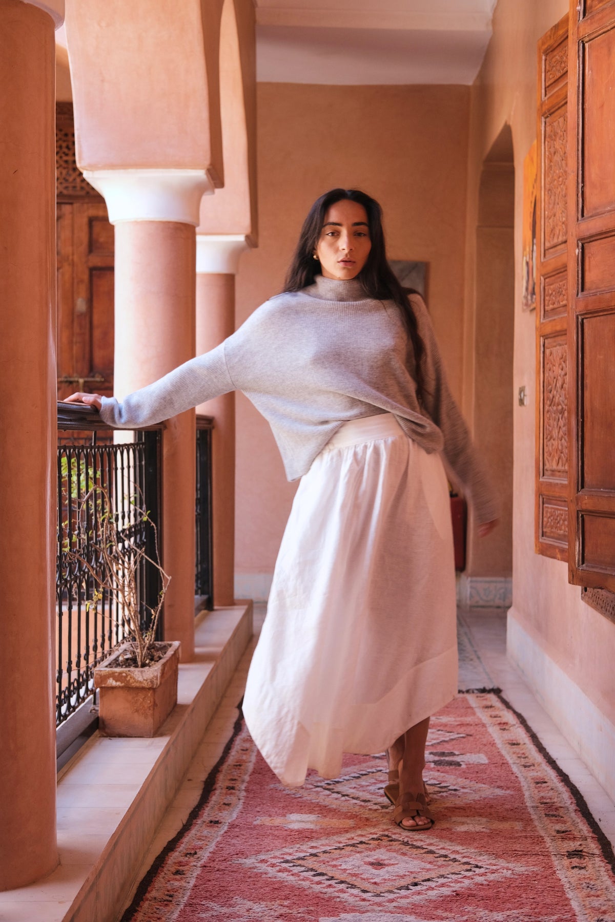 WomanLILLY PILLY woman wearing a grey Lumi cashmere knit sweater and ivory white Hannah skirt  standing in a hallway with columns and wooden doors