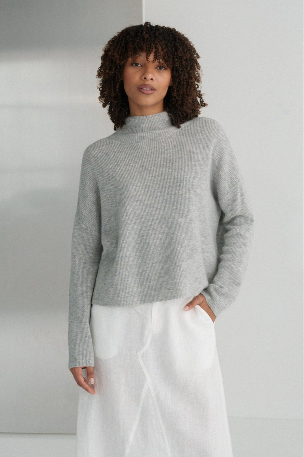 LILLY PILLY woman wearing a grey Lumi cashmere knit sweater and white Gia skirt standing in a minimalistic room.