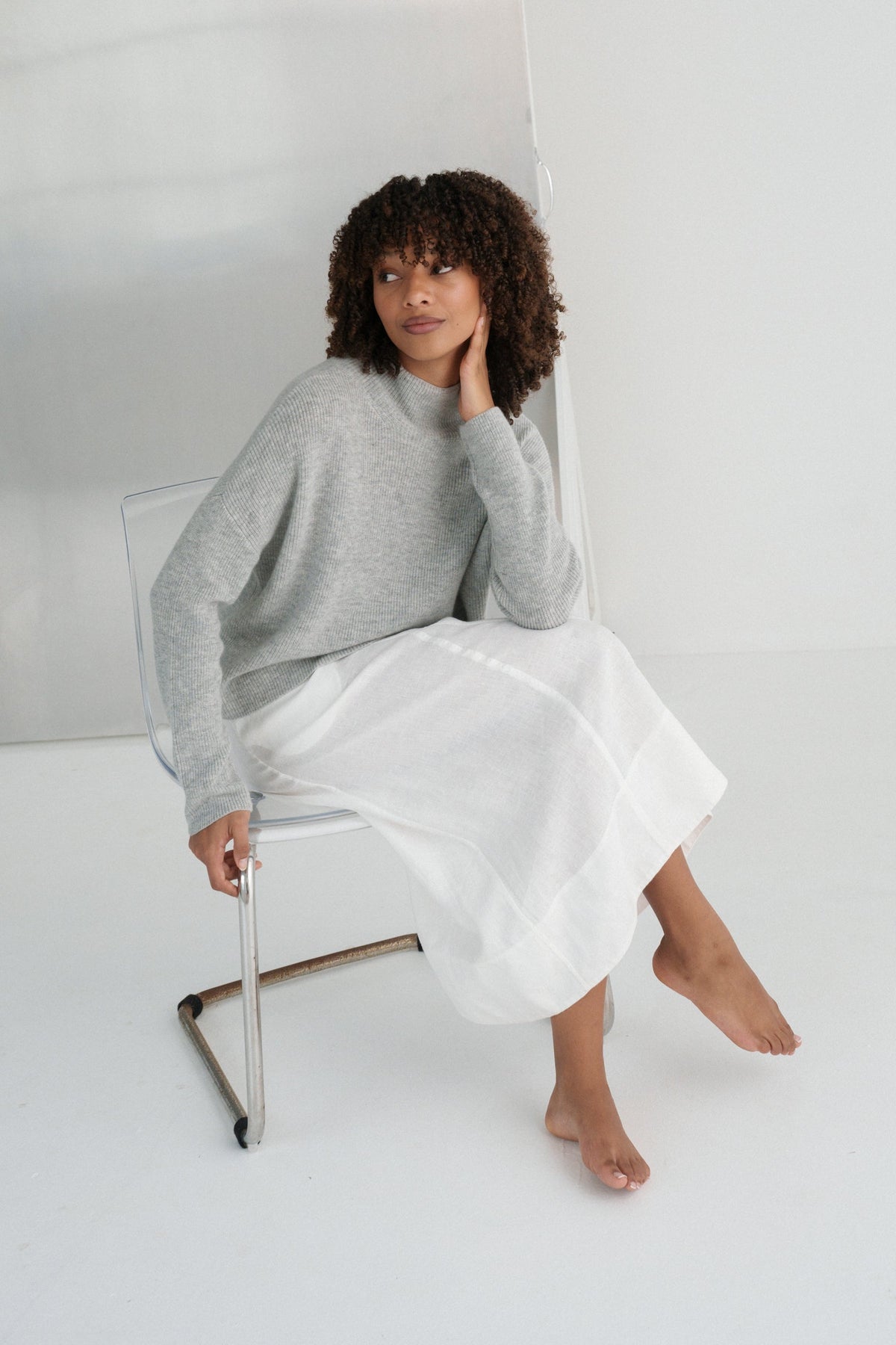LILLY PILLY woman wearing a grey Lumi cashmere knit sweater and white Gia skirt sitting on a chair