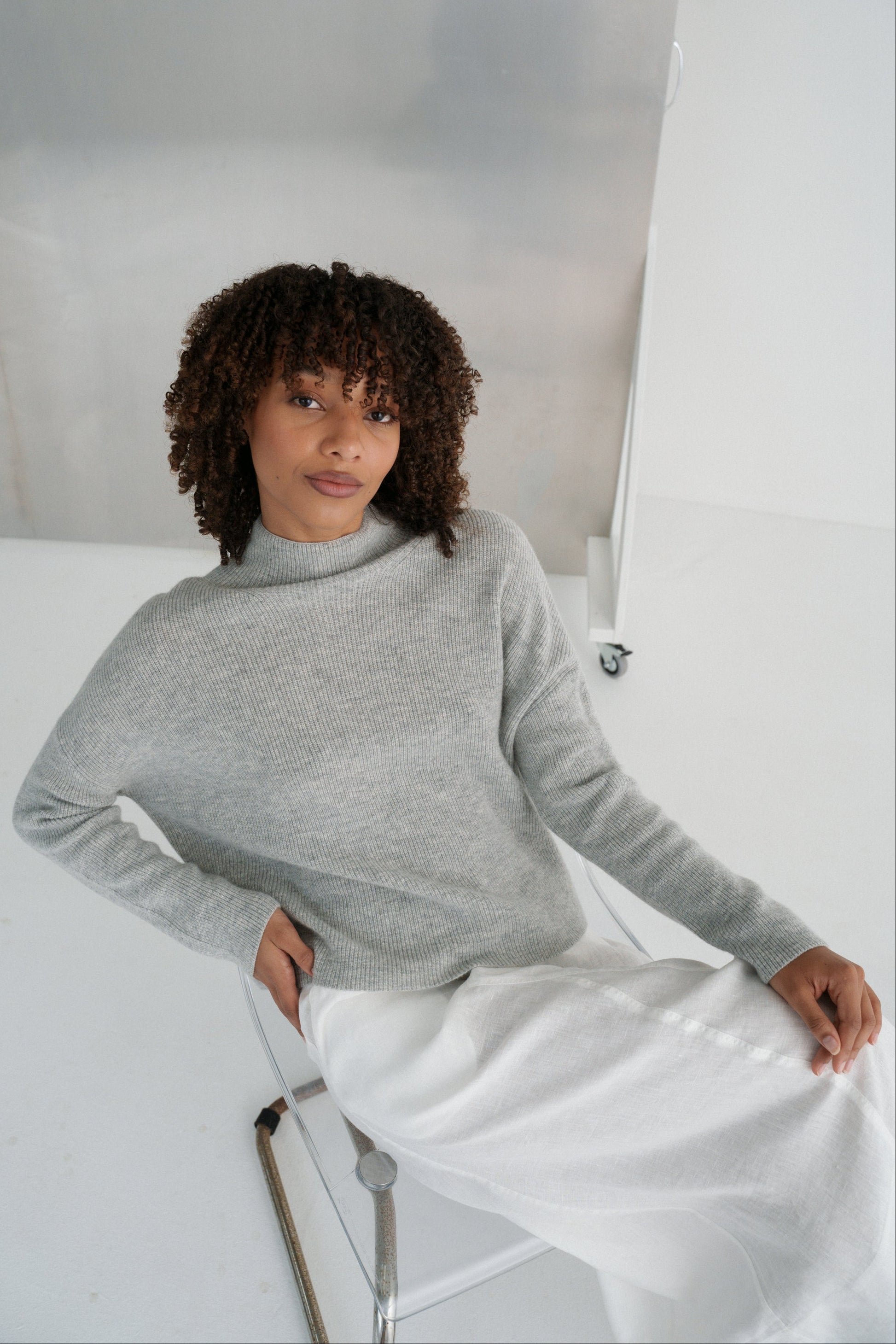 LILLY PILLY woman wearing a grey Lumi cashmere knit sweater and ivory white Gia skirt sitting on a white chair against a light background