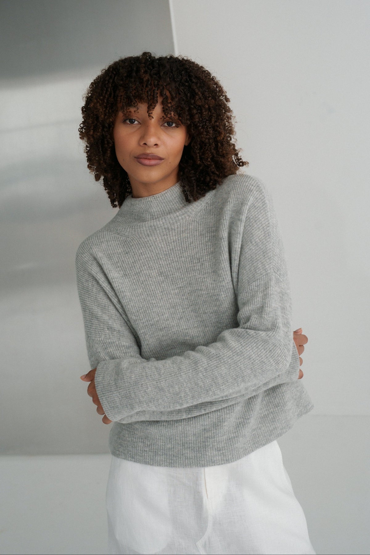 LILLY PILLY woman wearing a grey Lumi cashmere knit sweater and ivory white Gia skirt  against a plain background with arms folded
