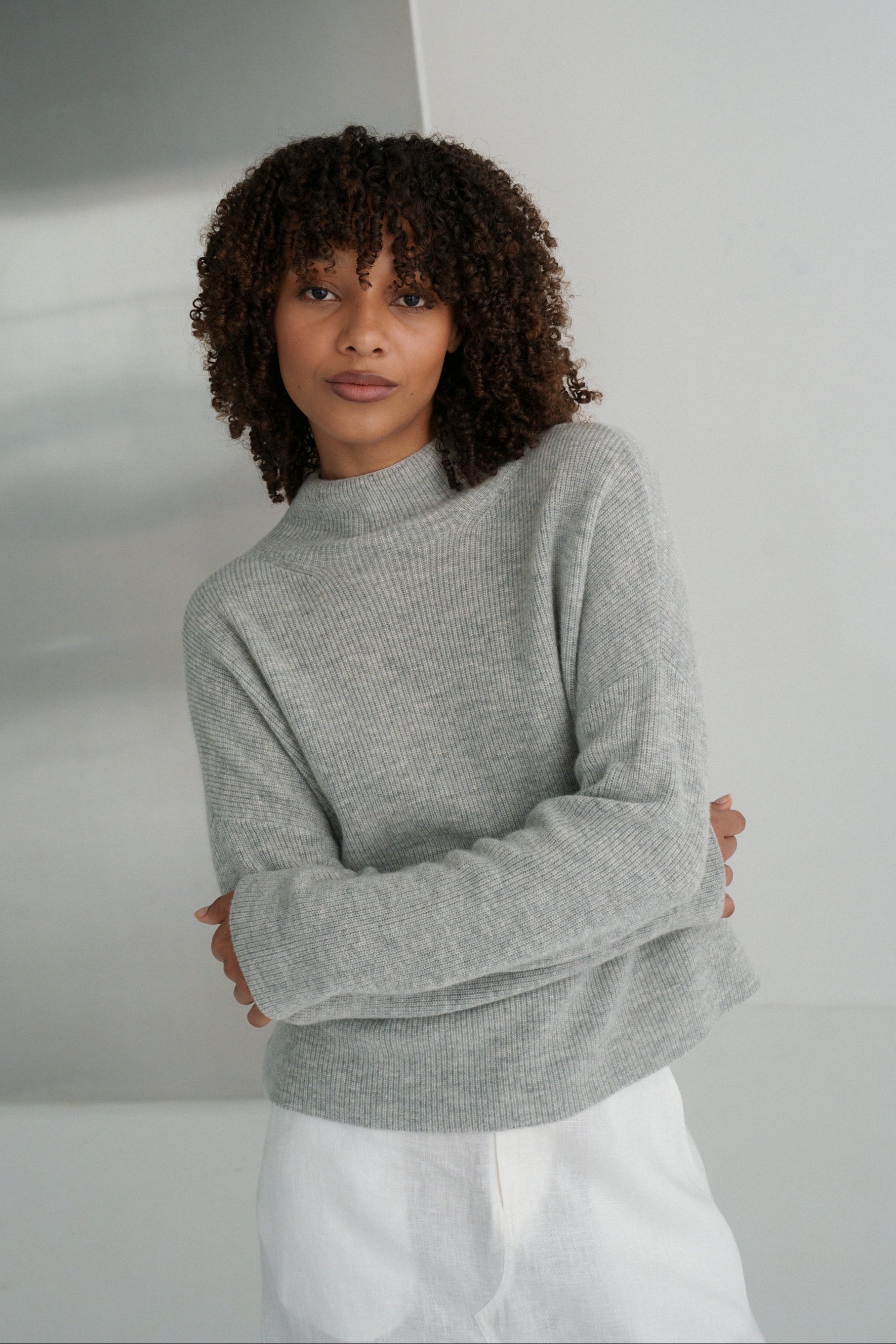 LILLY PILLY woman wearing a grey Lumi cashmere knit sweater and ivory white Gia skirt  against a plain background with arms folded