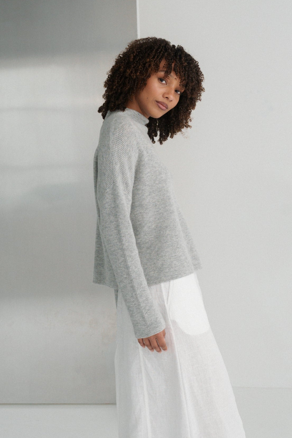 Side view of LILLY PILLY woman wearing a grey Lumi cashmere knit sweater and white Gia skirt standing against a white wall.