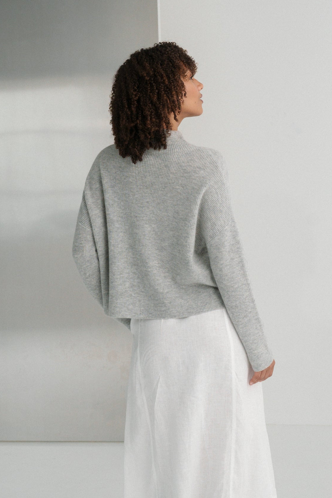 Back view of LILLY PILLY woman wearing a grey Lumi cashmere knit sweater and white Gia skirt standing in a minimalistic room.