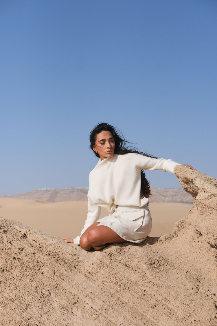 Lumi Cashmere Knit in Ivory - front view, seated on sand under a clear blue sky - Lilly Pilly Collection