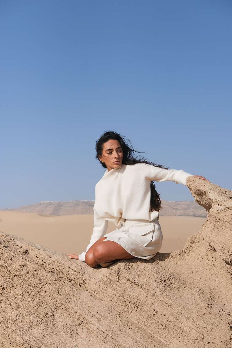 LILLY PILLY woman in ivory Lumi cashmere knit and Betty linen skirt sitting on a rock with a clear blue sky