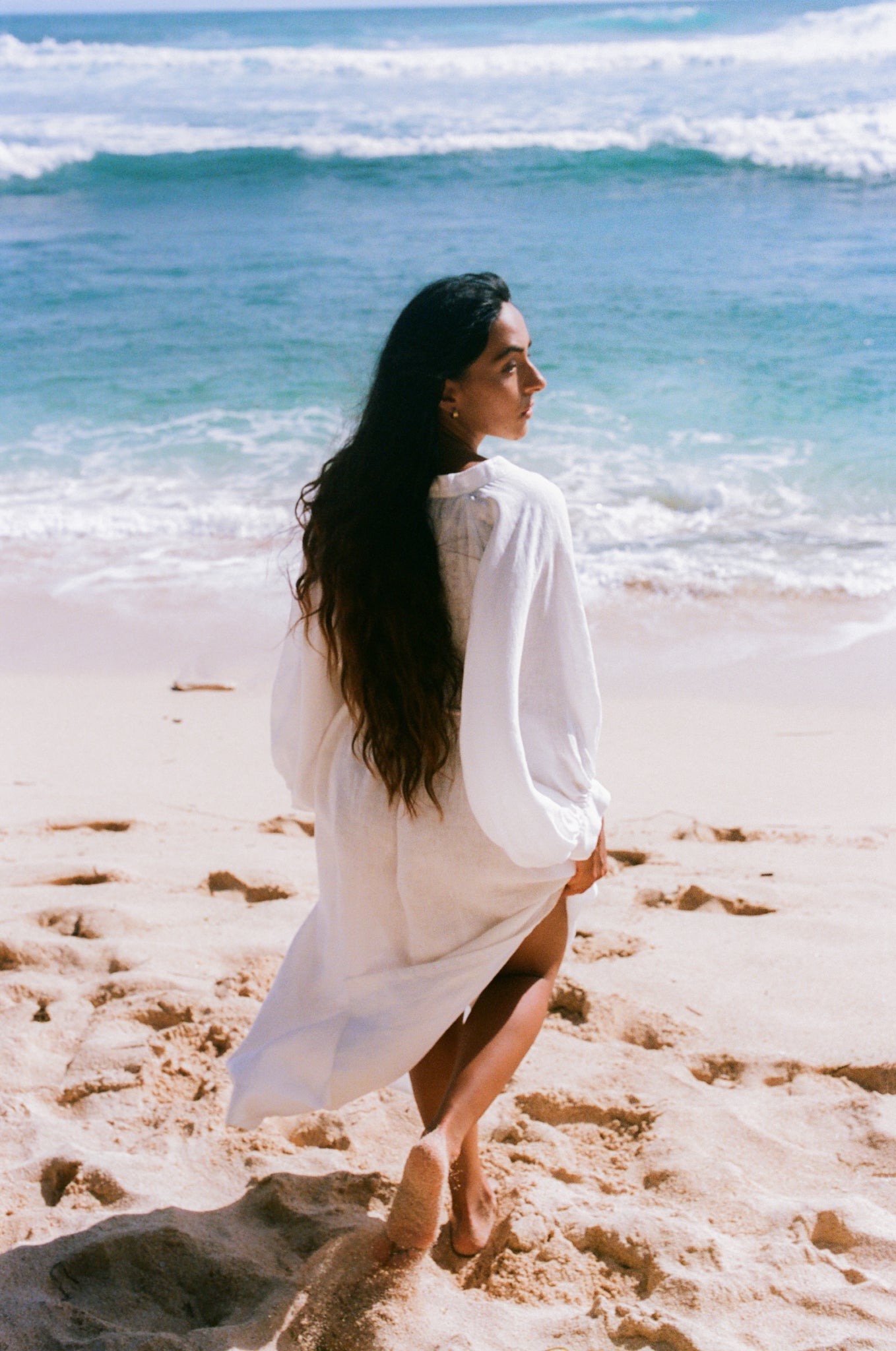Woman in an ivory Mae linen dress standing on a beach with ocean waves in the background back view
