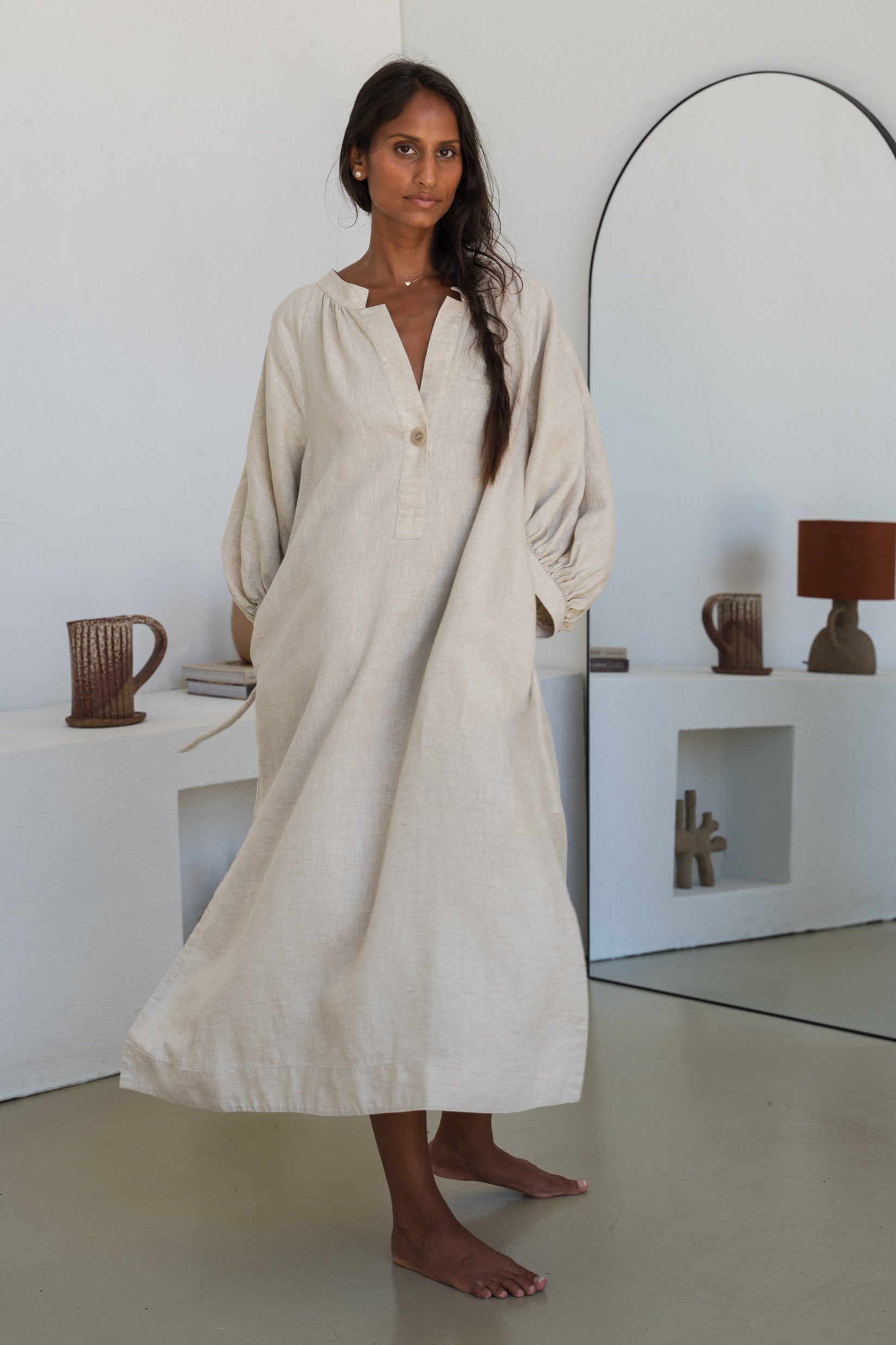 Woman wearing a Mae oatmeal linen dress twirling in a room with white walls and a mirror.