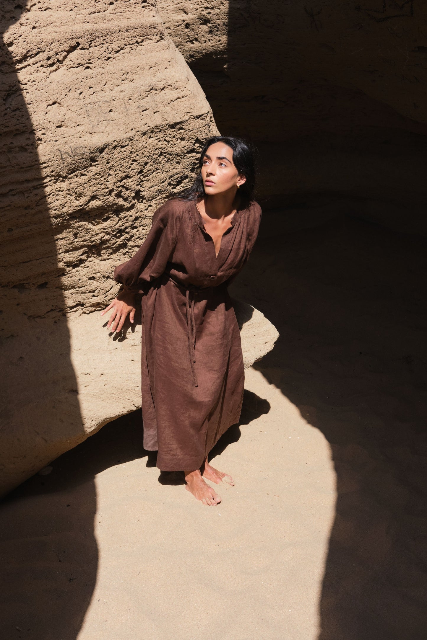 LILLY PILLY woman wearing a chocolate brown Mae linen dress standing against a textured wall with a dramatic light effect.