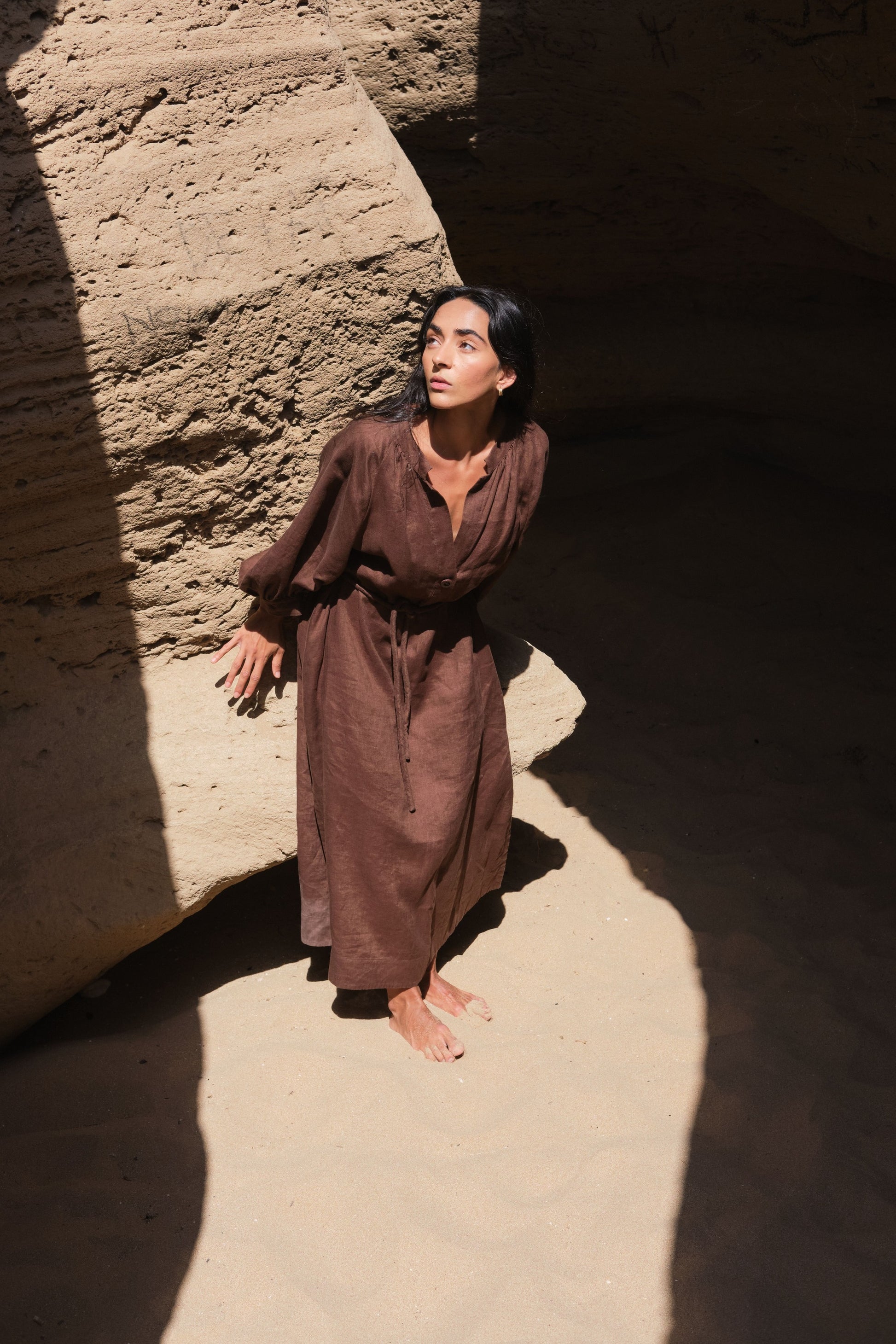 LILLY PILLY woman wearing a chocolate brown Mae linen dress standing against a textured wall with a dramatic light effect.
