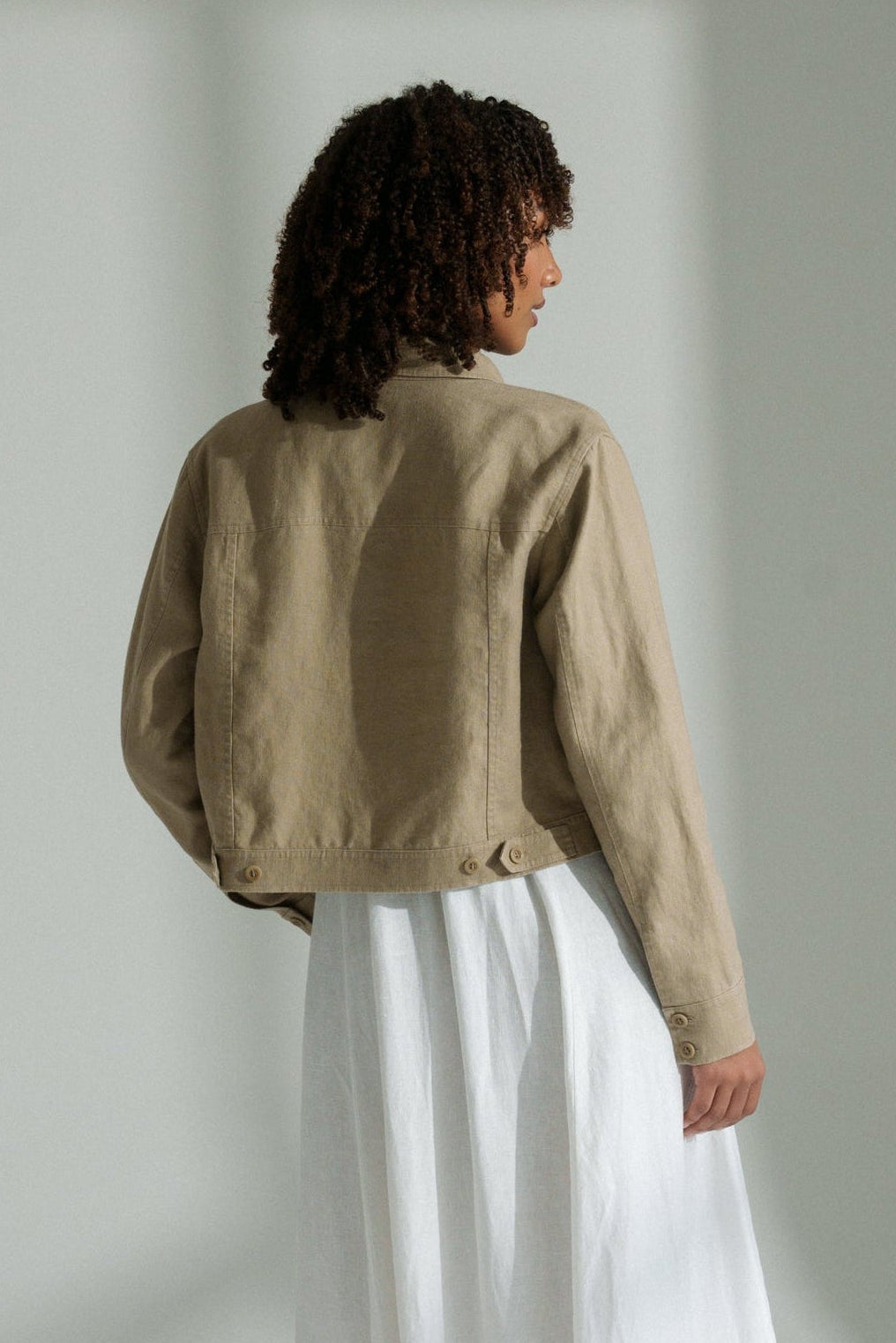 LILLY PILLY woman wearing a taupe Milly linen jacket and white pants standing against a white wall.