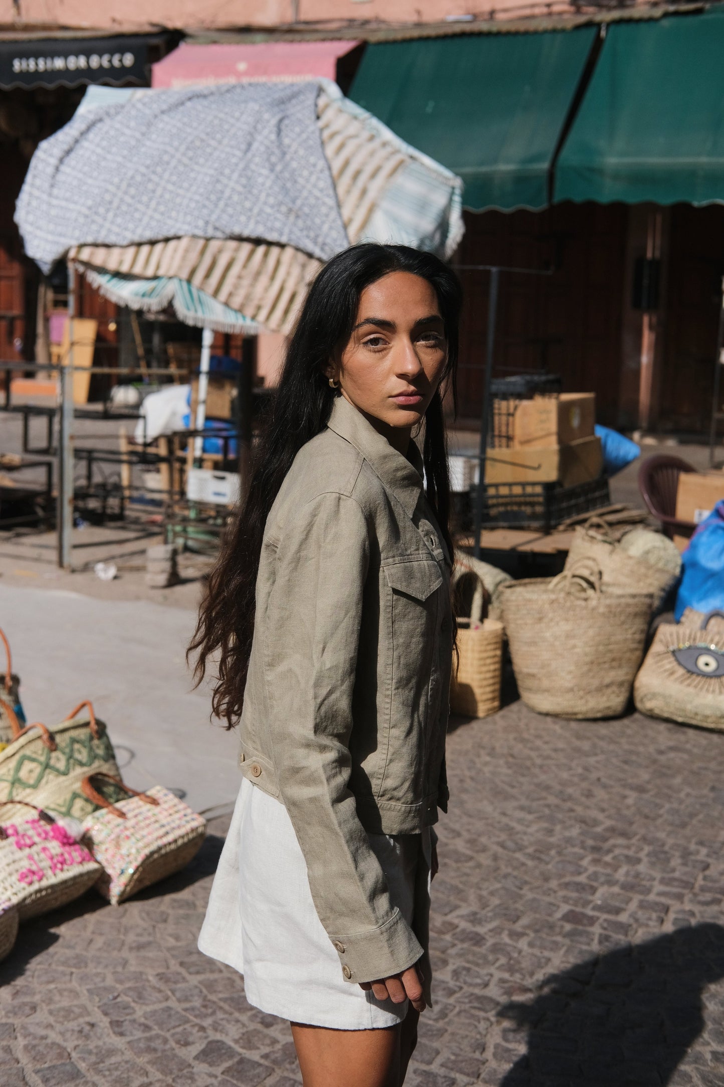 LILLY PILLY woman wearing a taupe Milly linen jacket standing in an outdoor market setting with various items and a green canopy in the background.