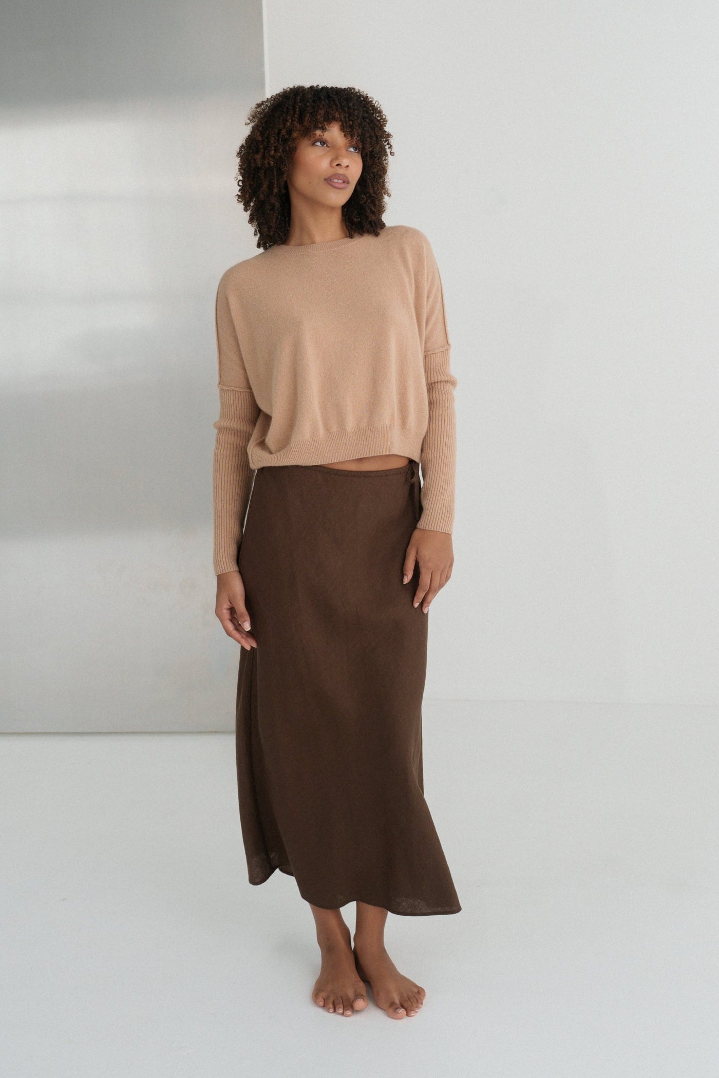 LILLY PILLY woman wearing Camel brown Miri cashmere knit sweater standing against a white wall.
