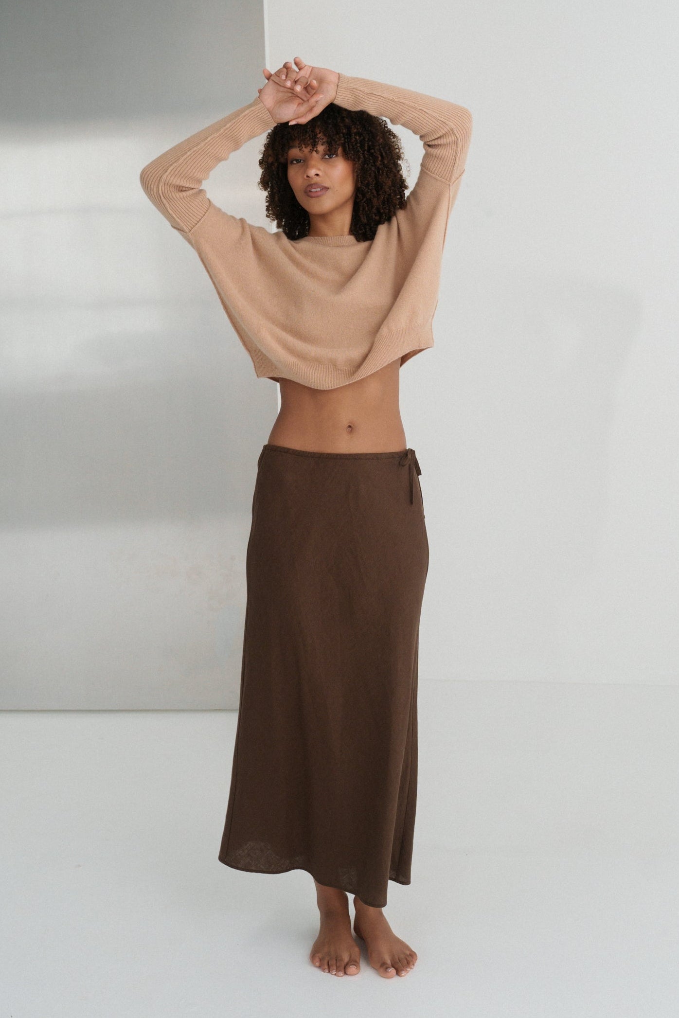LILLY PILLY woman wearing Camel brown Miri cashmere knit sweater  standing against a white wall.