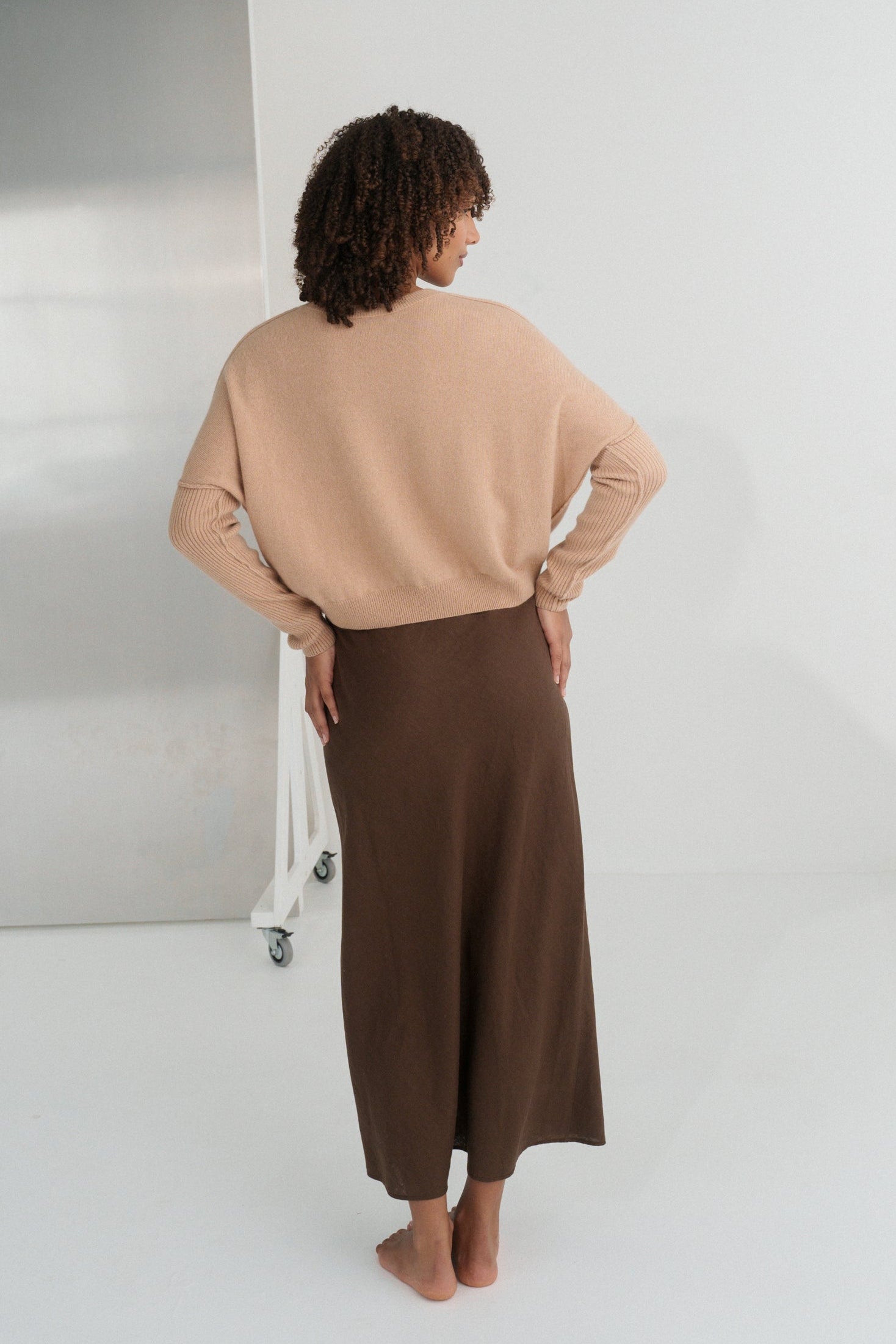 Back view of LILLY PILLY woman wearing Camel brown Miri cashmere knit sweater  standing in a minimalistic room with a mirror.