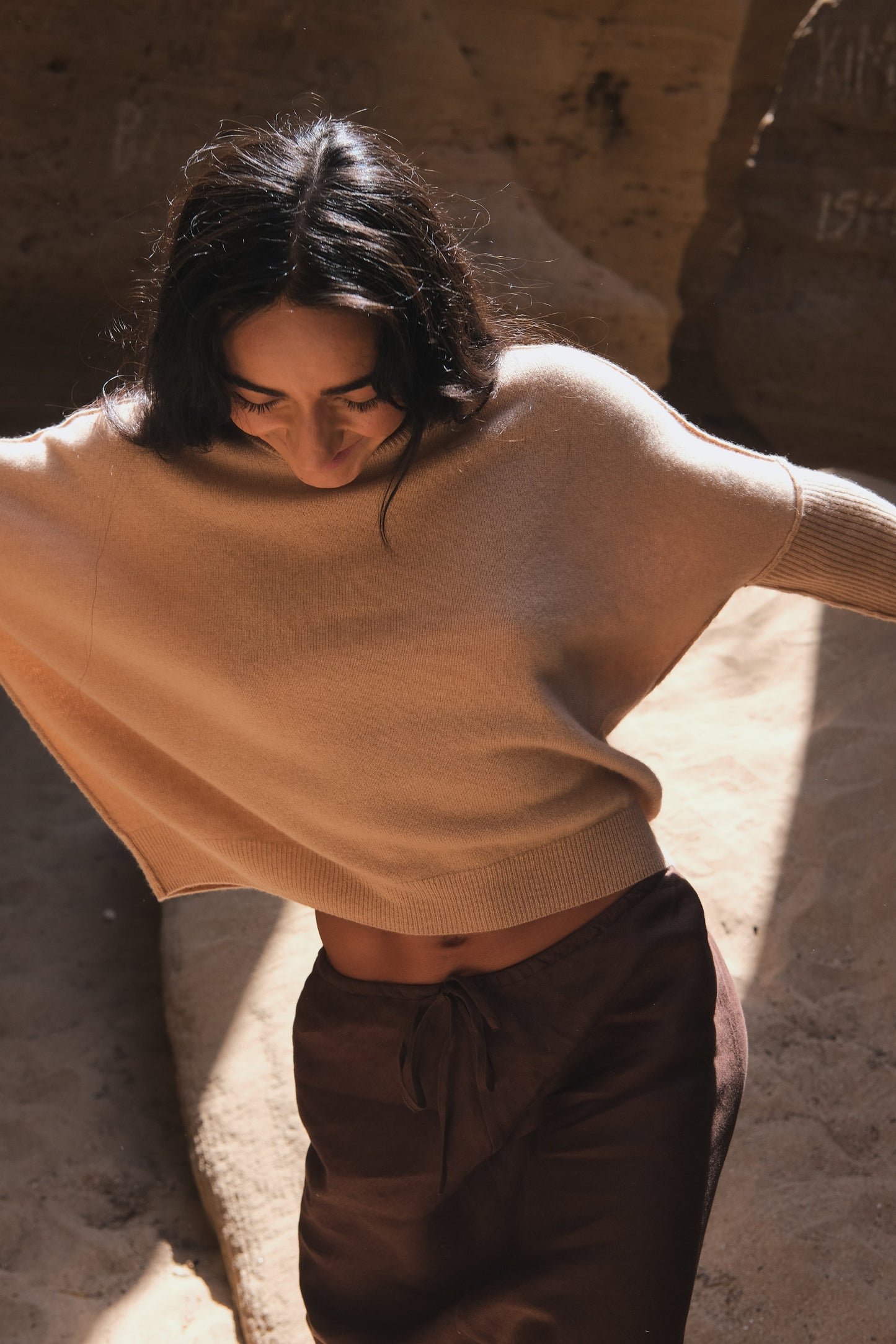 LILLY PILLY woman wearing Camel brown Miri cashmere knit sweater and chocolate brown skirt against a textured wall.