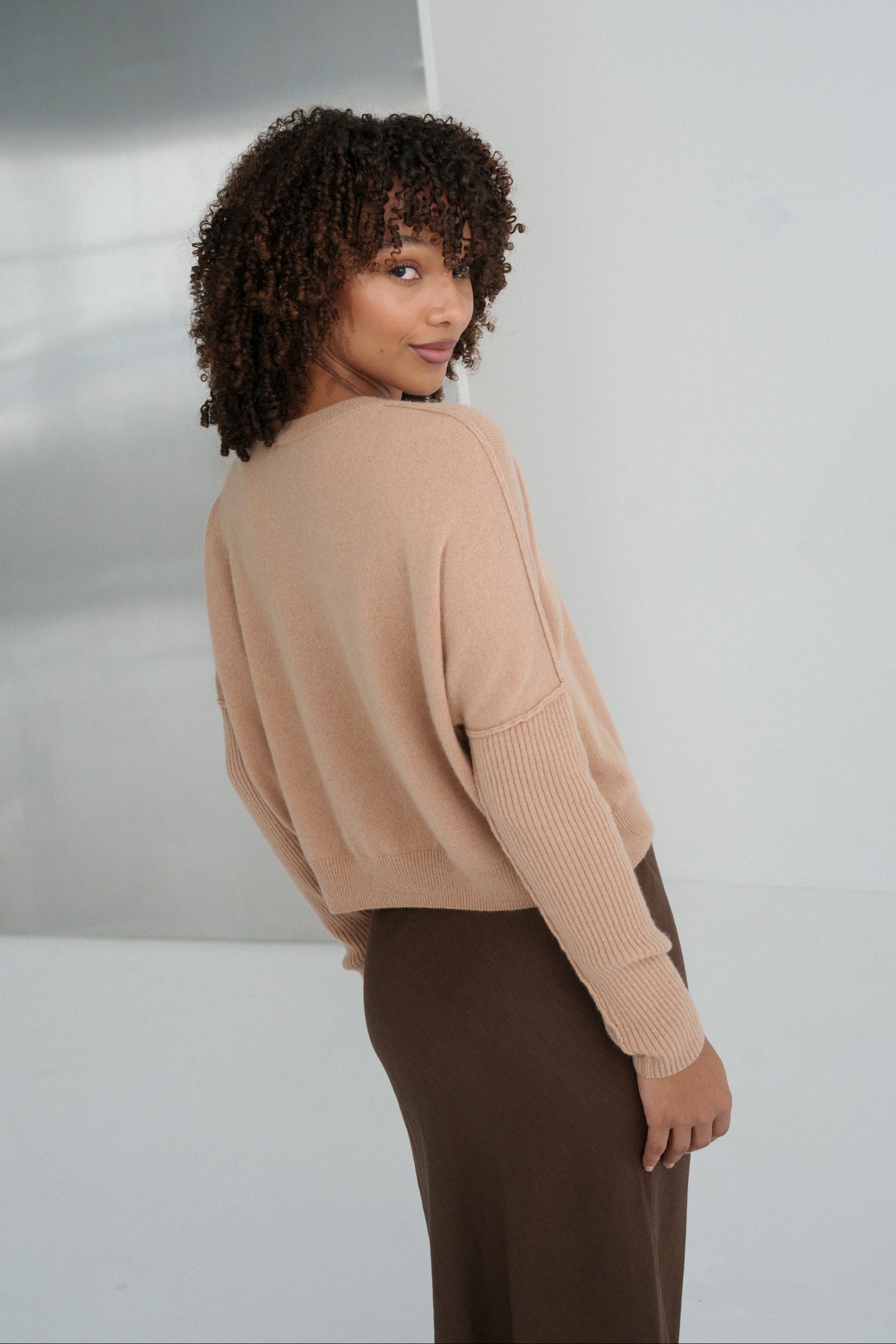 Back view of LILLY PILLY woman wearing Camel brown Miri cashmere knit sweater looking over her shoulder while standing against a white wall.