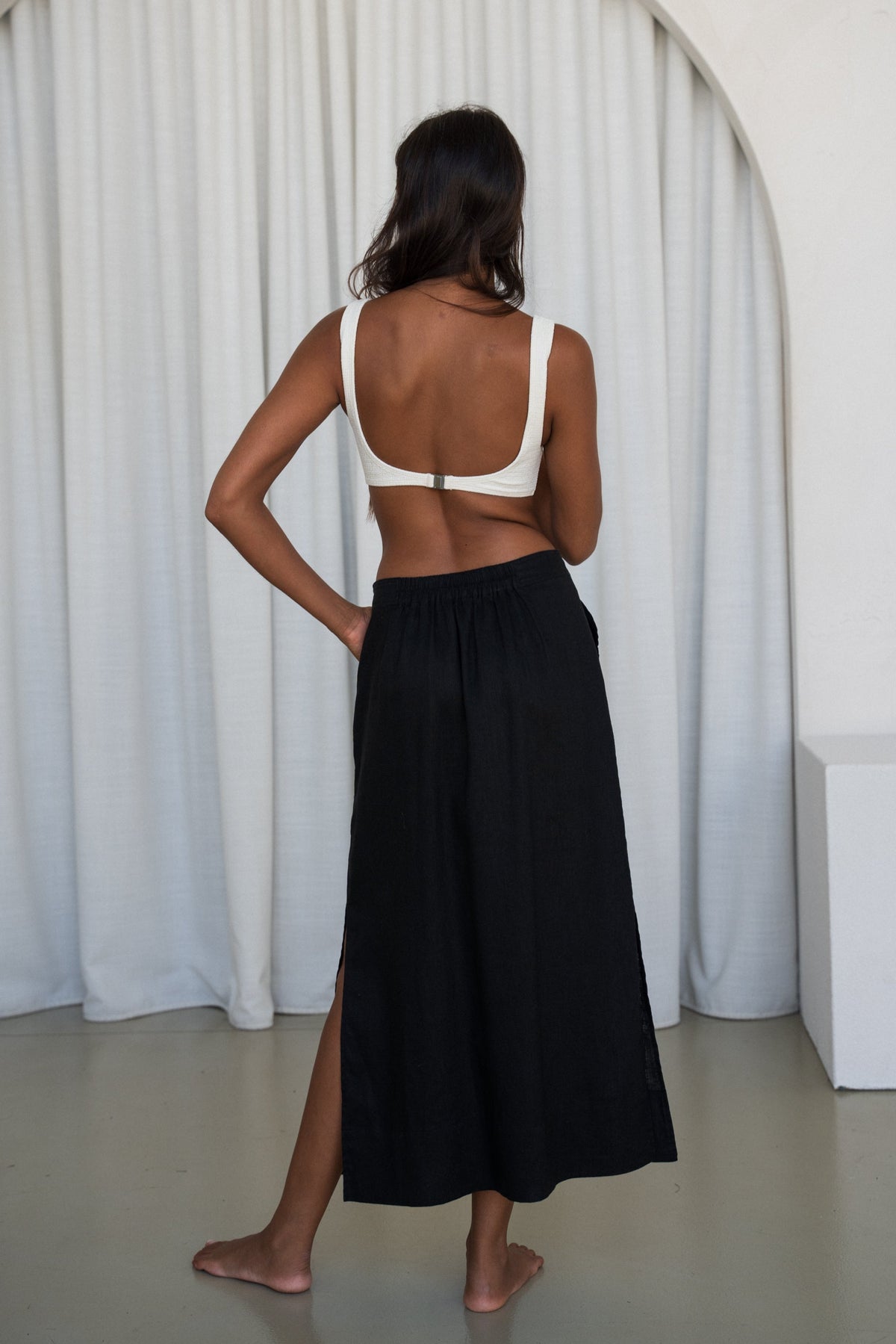 Back view of woman wearing a white top and black Nadi linen skirt standing against a white curtain.