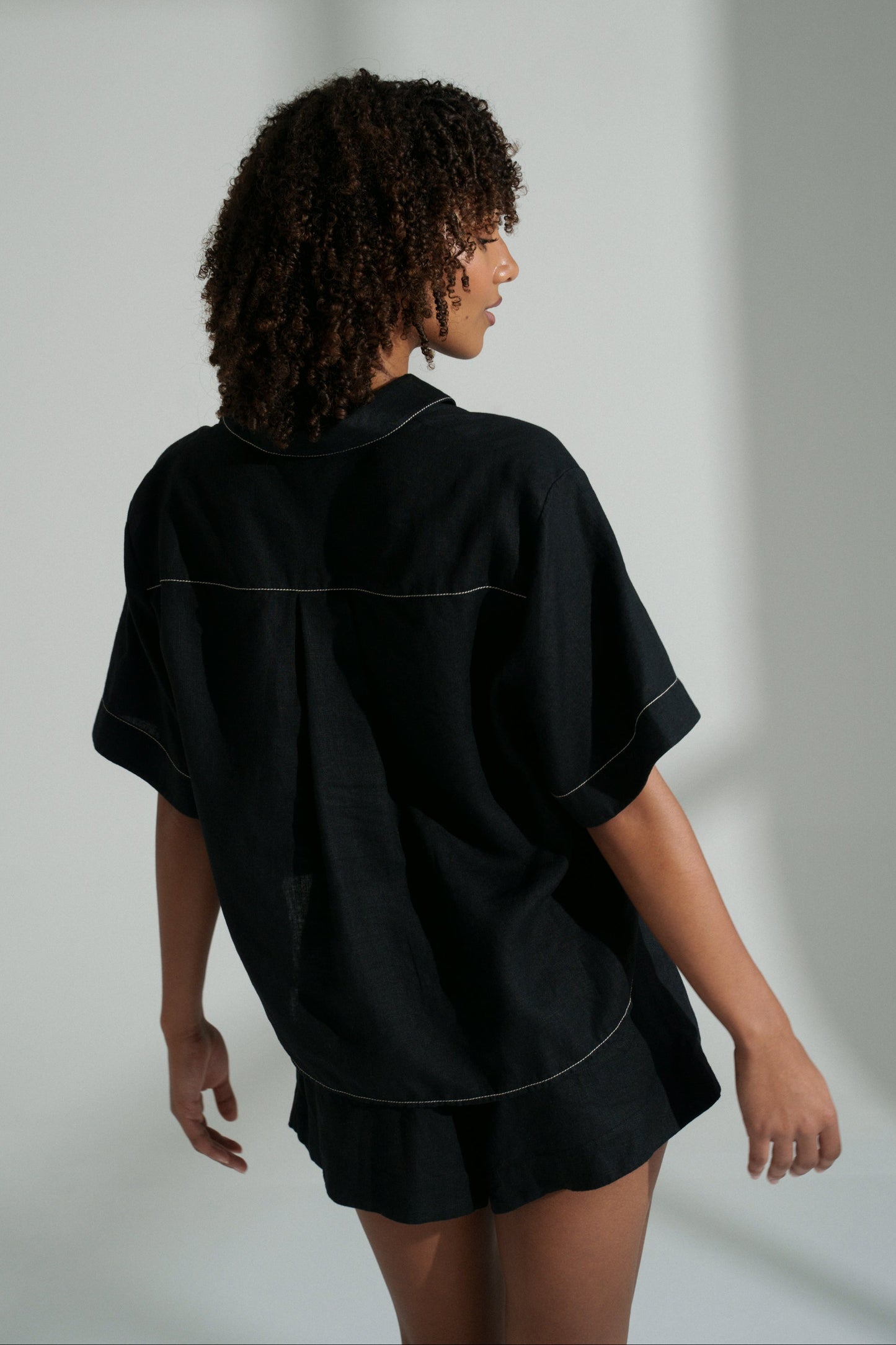 LILLY PILLY woman wearing a black Nika linen shirt top and Gaia linen shorts set against a white background showing back view