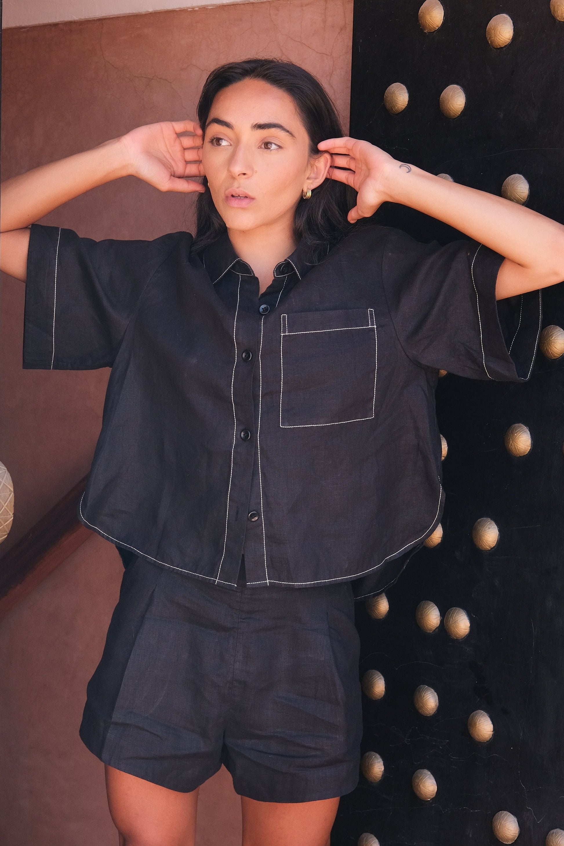 LILLY PILLY woman wearing a black Nika linen shirt top and Gaia linen shorts outfit standing in front of a large ornate door.