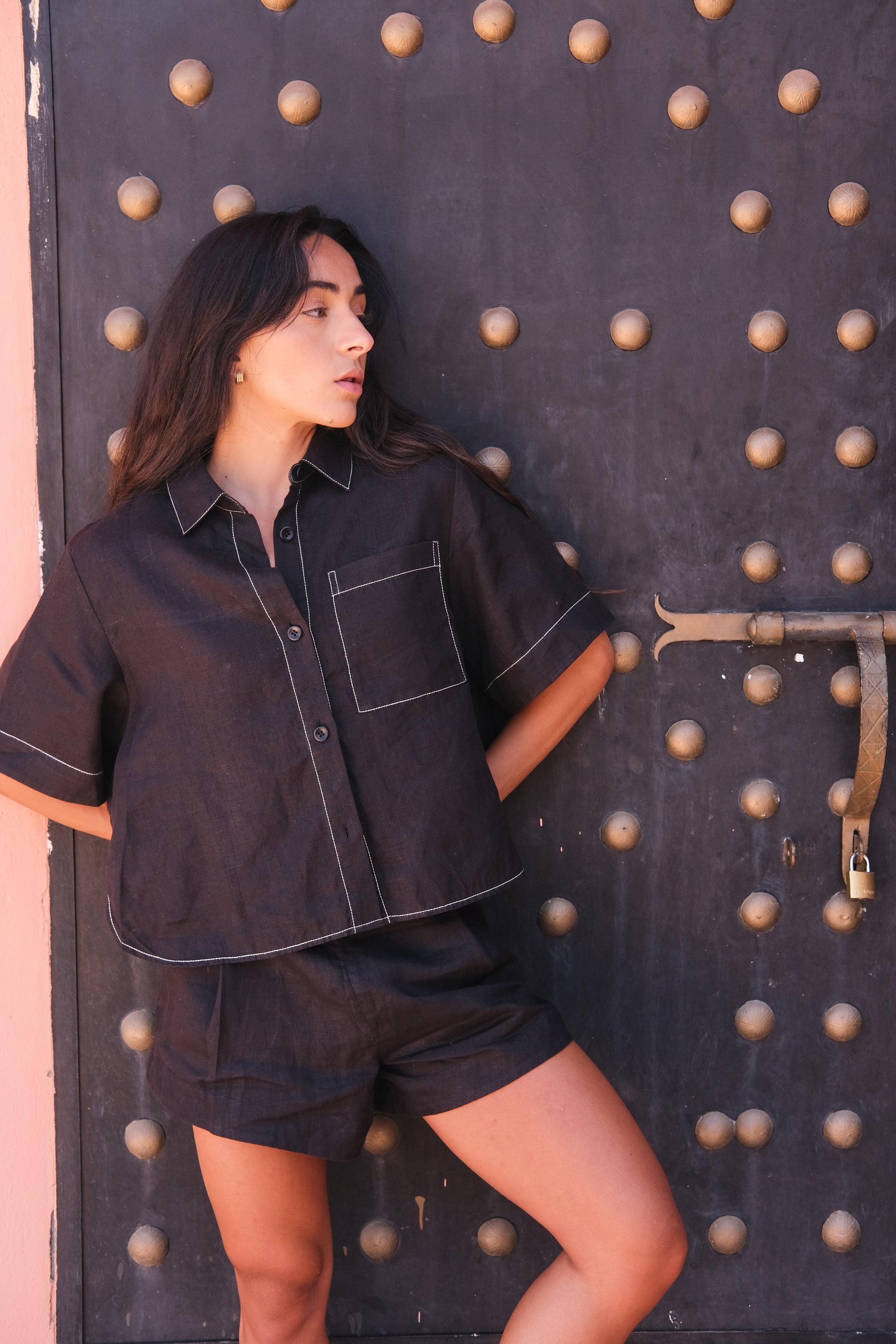 LILLY PILLY woman wearing a black Nika linen shirt top and Gaia linen shorts outfit against against a textured wall