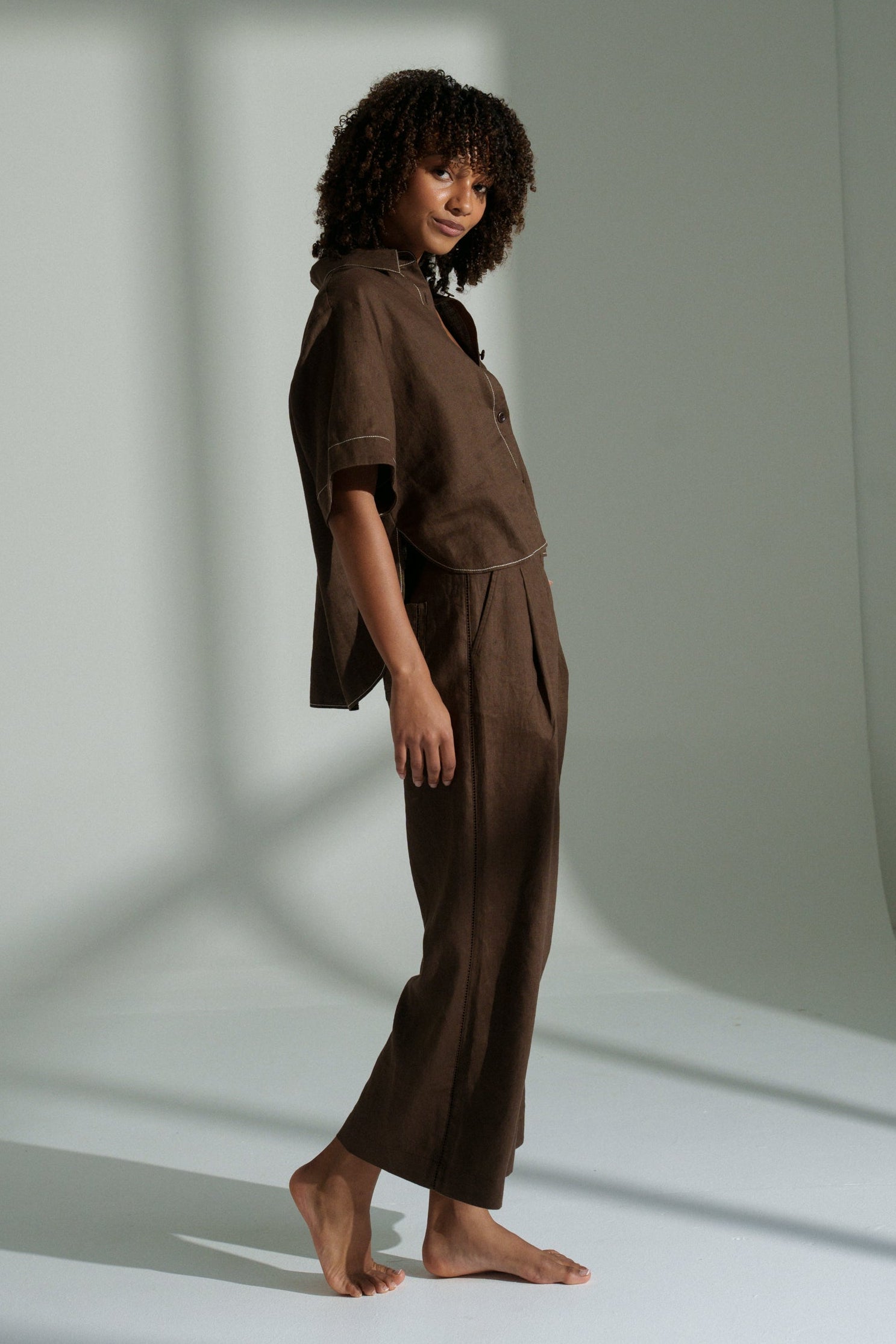 LILLY PILLY woman wearing a chocolate Nika linen shirt top standing in a room with light casting shadows on the wall.