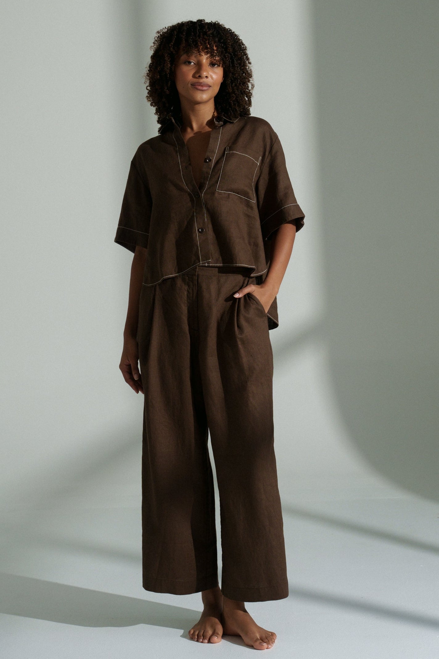 LILLY PILLY woman wearing a chocolate brown Nika linen shirt top standing against a white wall with shadow patterns.
