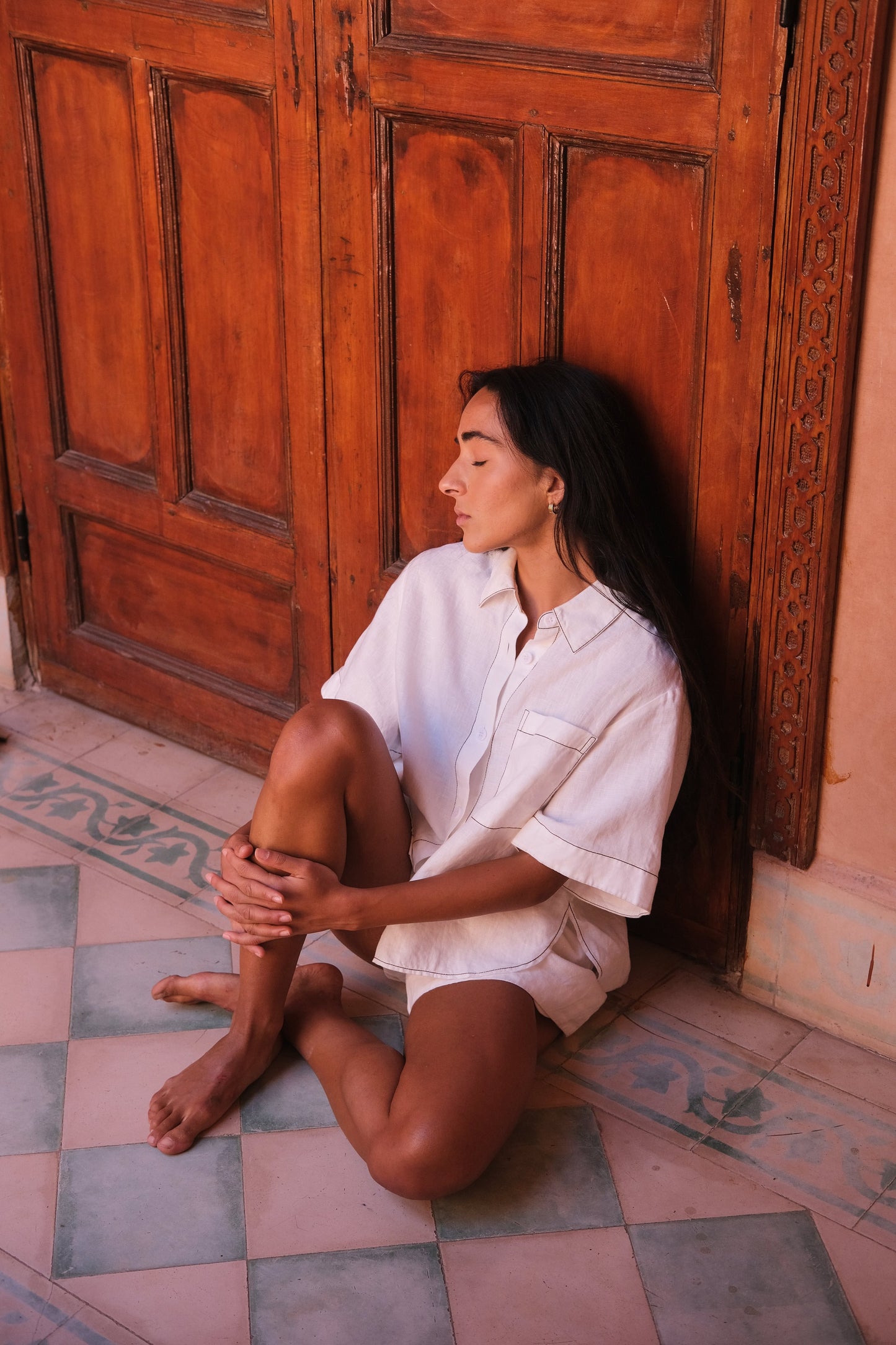 LILLY PILLY woman wearing an ivory Nika linen shirt top and Gaia linen shorts outfit sitting against a wooden door.