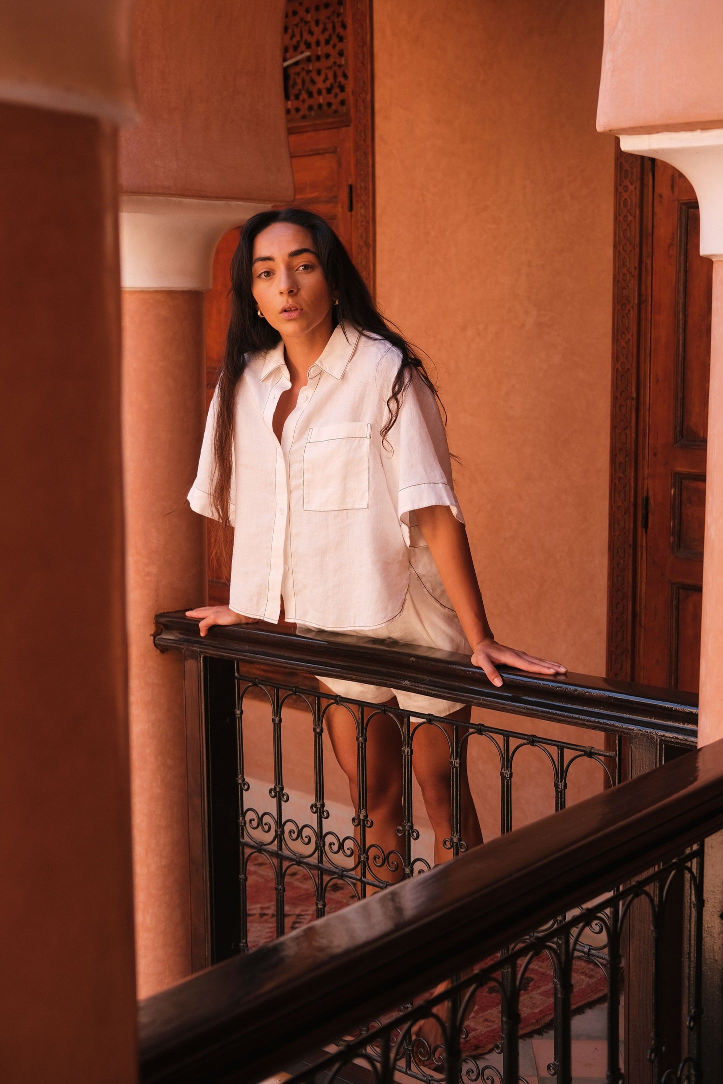 LILLY PILLY woman wearing an ivory Nika linen shirt top and Gaia linen shorts outfit standing on a balcony with warm-toned walls.
