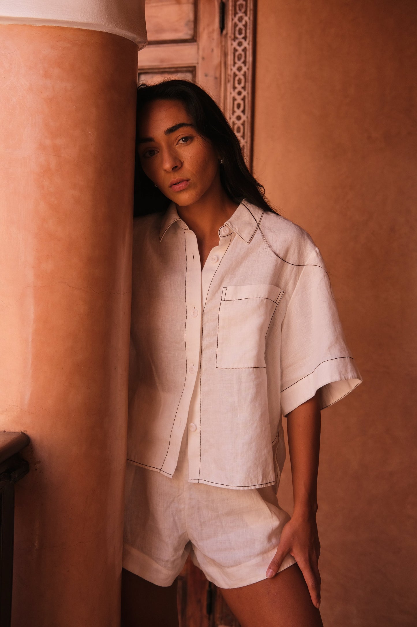 LILLY PILLY woman wearing an ivory Nika linen shirt top and Gaia linen shorts outfit standing against a warm-toned wall.