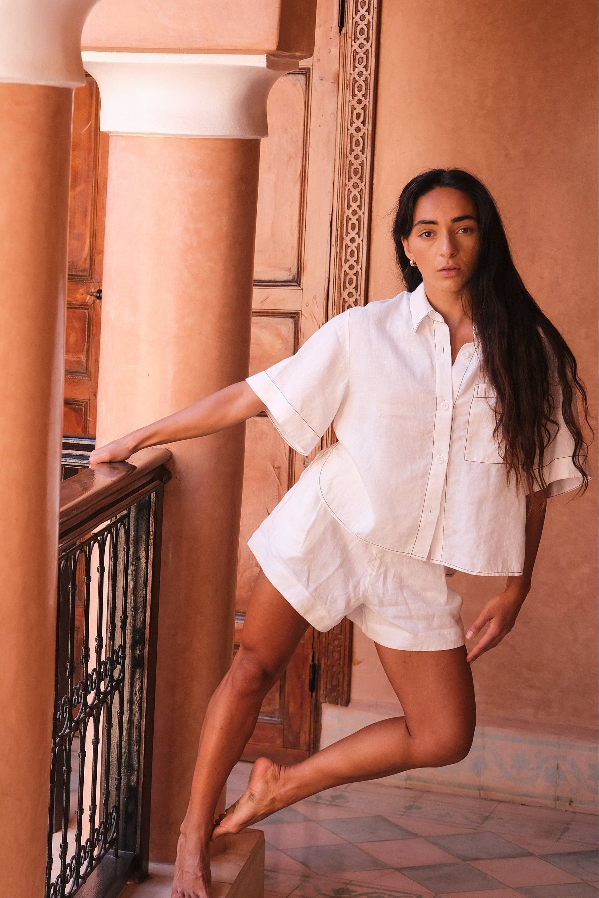LILLY PILLY woman wearing an ivory Nika linen shirt top and Gaia linen shorts outfit standing on a balcony with columns and a warm-toned wall.
