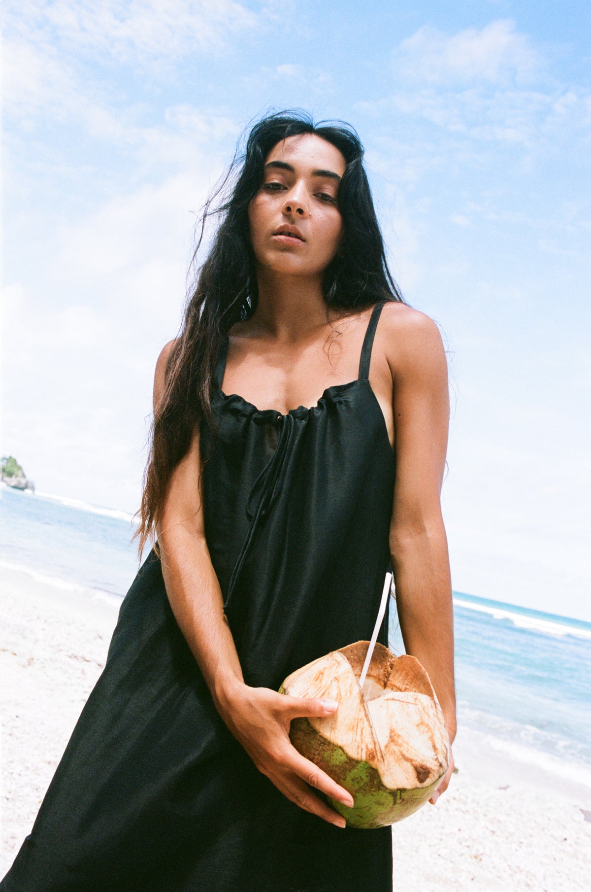 Woman in a black Nova silk dress holding a coconut on a beach