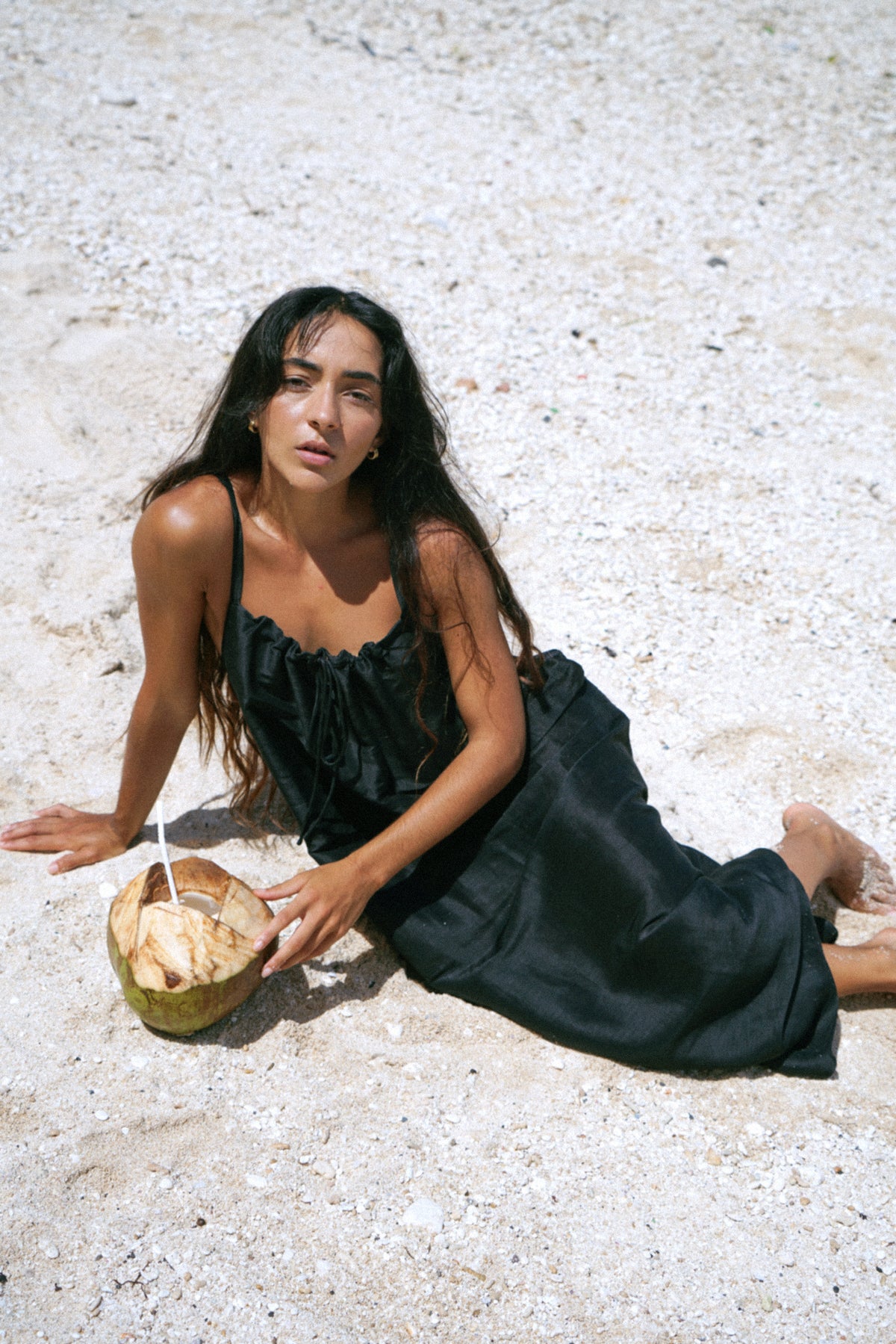 Woman in a black dress sitting on a sandy beach with a coconut