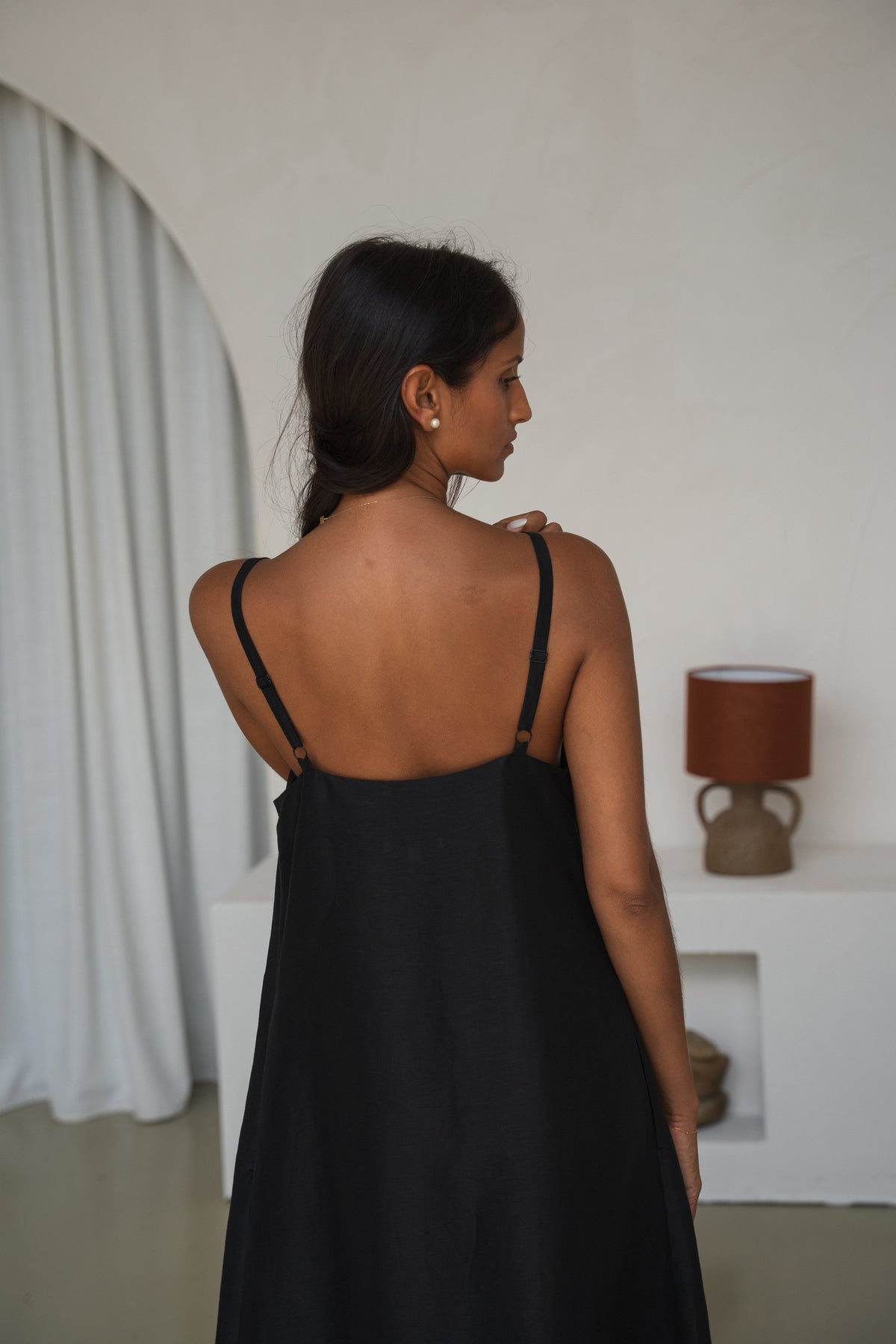 Back view of woman wearing a black slip dress in a room with white walls and a lamp.