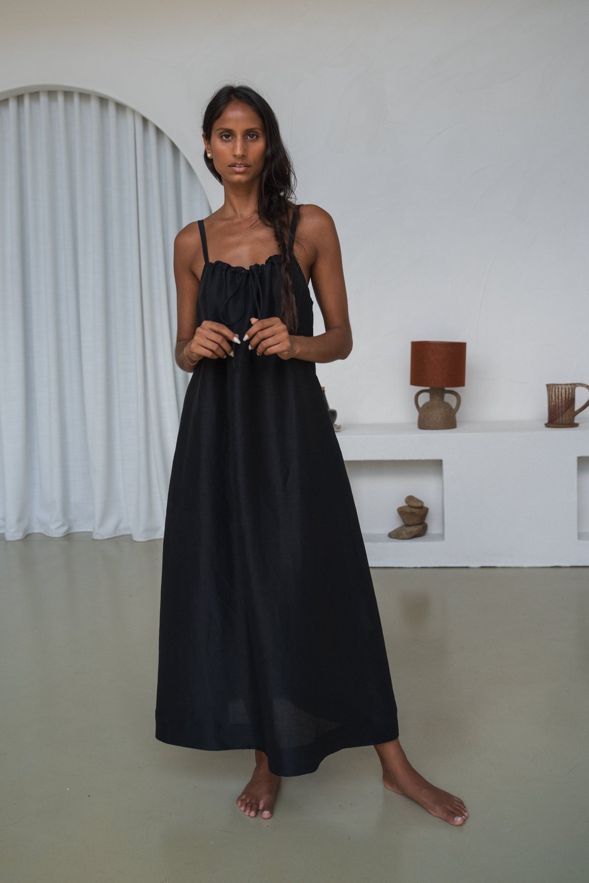 Woman wearing a black dress standing in a minimalistic room with white walls and a white shelf.