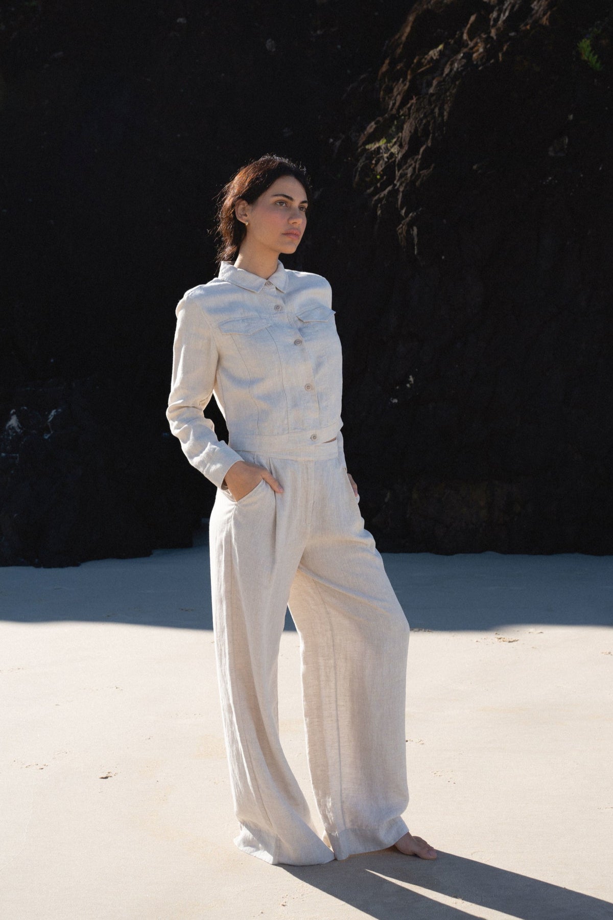 LILLY PILLY woman wearing a Milly linen jacket and Oli linen pants in oatmeal standing on a sandy beach with dark cliffs in the background