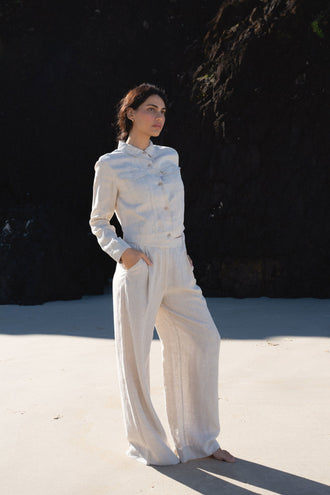 Oli Linen Pants in Oatmeal - three-quarter side view, woman wearing Oatmeal wide-leg linen pants with matching Milly linen jacket, hand in pocket, sandy beach with dark cliff backdrop - Lilly Pilly Collection