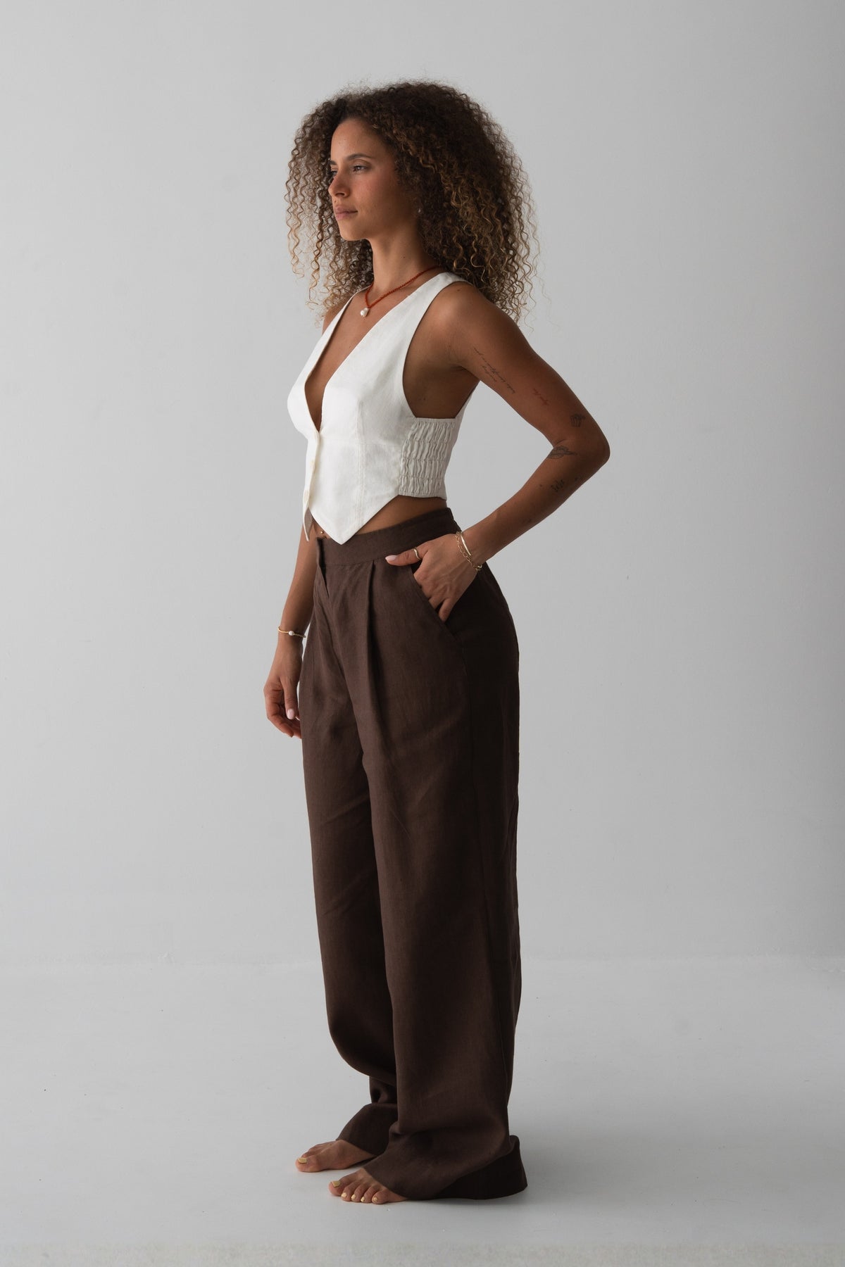 Oli Linen Pants in Chocolate - side view, woman wearing Chocolate wide-leg linen pants with ivory V-neck linen waistcoat, hand in pocket, studio backdrop - Lilly Pilly Collection