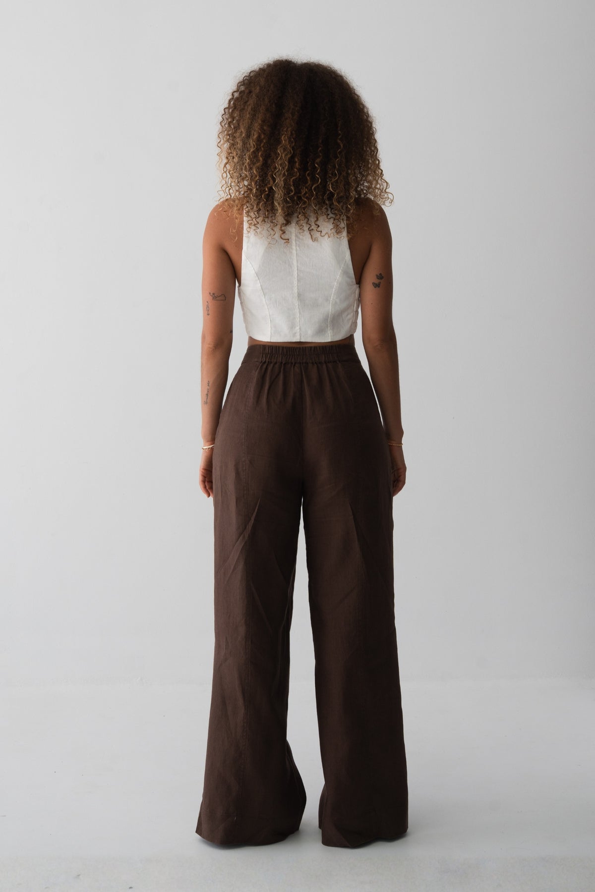 Oli Linen Pants in Chocolate - back view showing elasticated waistband and wide-leg linen silhouette, woman wearing ivory linen waistcoat, studio backdrop - Lilly Pilly Collection