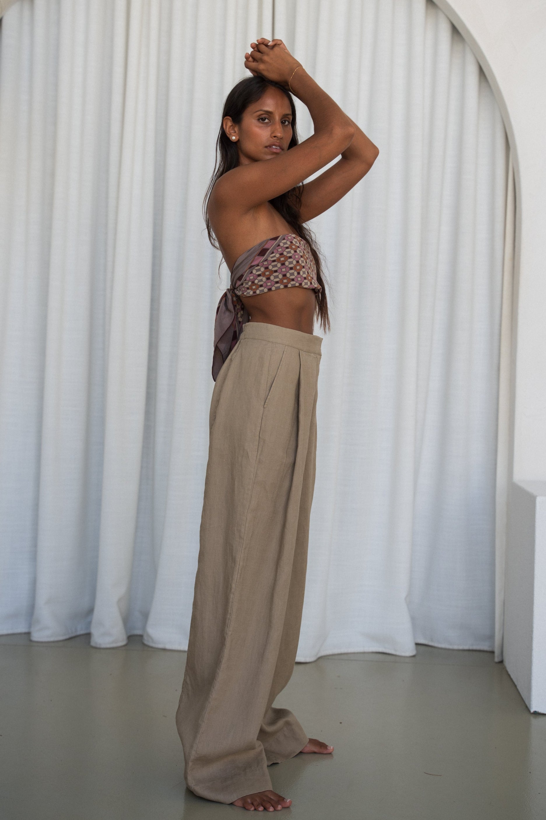 LILLY PILLY woman wearing Oli linen pants in Taupe, side view with white curtain background