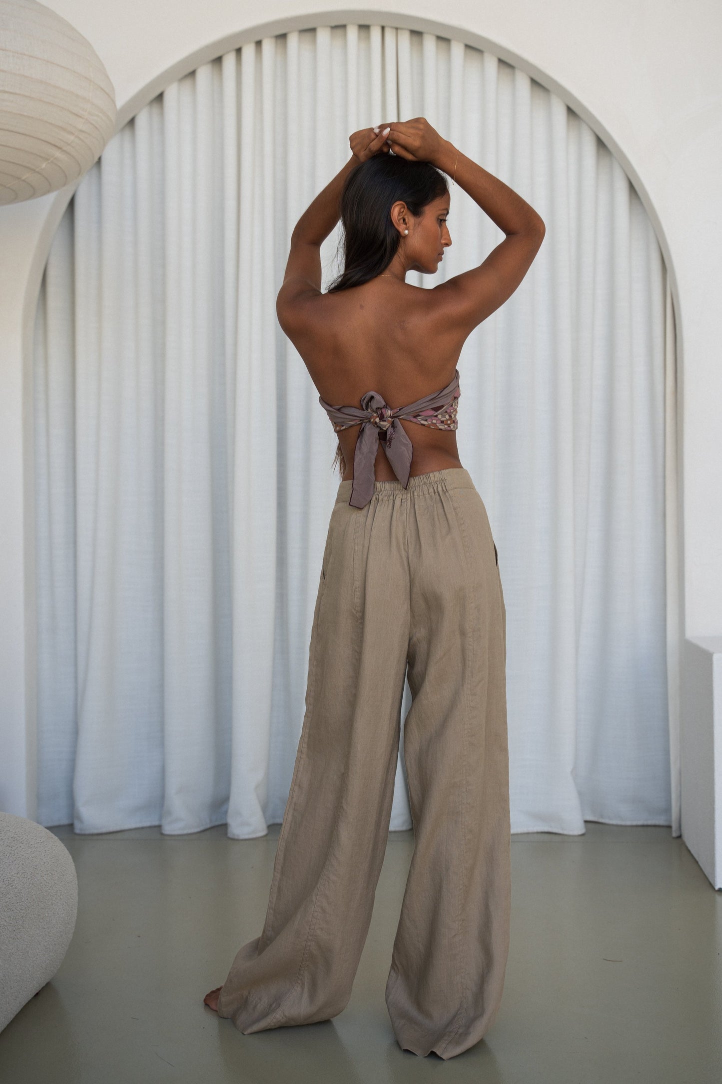 LILLY PILLY woman wearing Oli linen pants in Taupe, back view with white curtain background