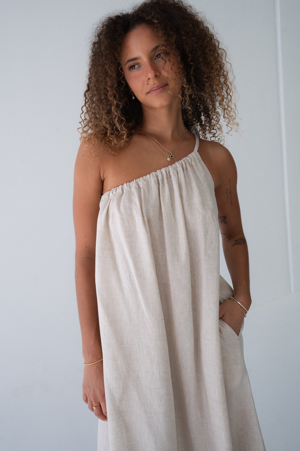 Ophelia Linen Midi Dress in Oatmeal - three-quarter view, asymmetric gathered neckline, hand in pocket - Lilly Pilly Collection