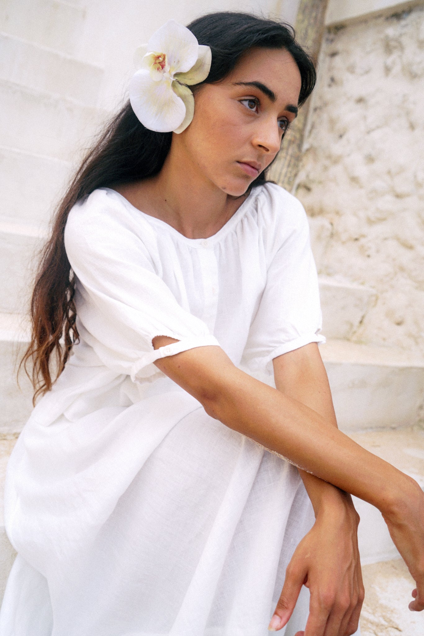 LILLY PILLY woman wearing an ivory Pia linen shirt top with a flower in her hair, sitting against a light stone wall.