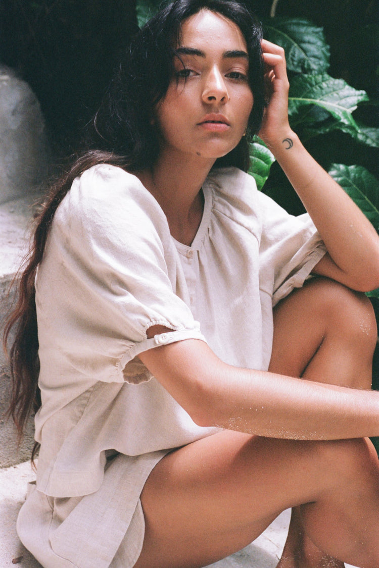 Pia Linen Top in Oatmeal - woman seated, round gathered neckline and puff sleeves, tropical backdrop - Lilly Pilly Collection