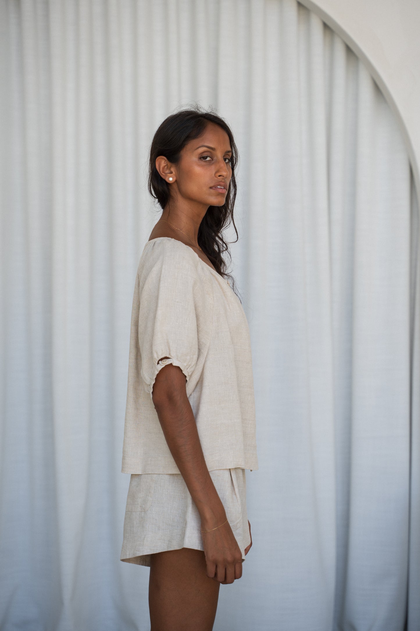 LILLY PILLY woman wearing a oatmeal Pia linen shirt top and Gaia shorts with a curtain background