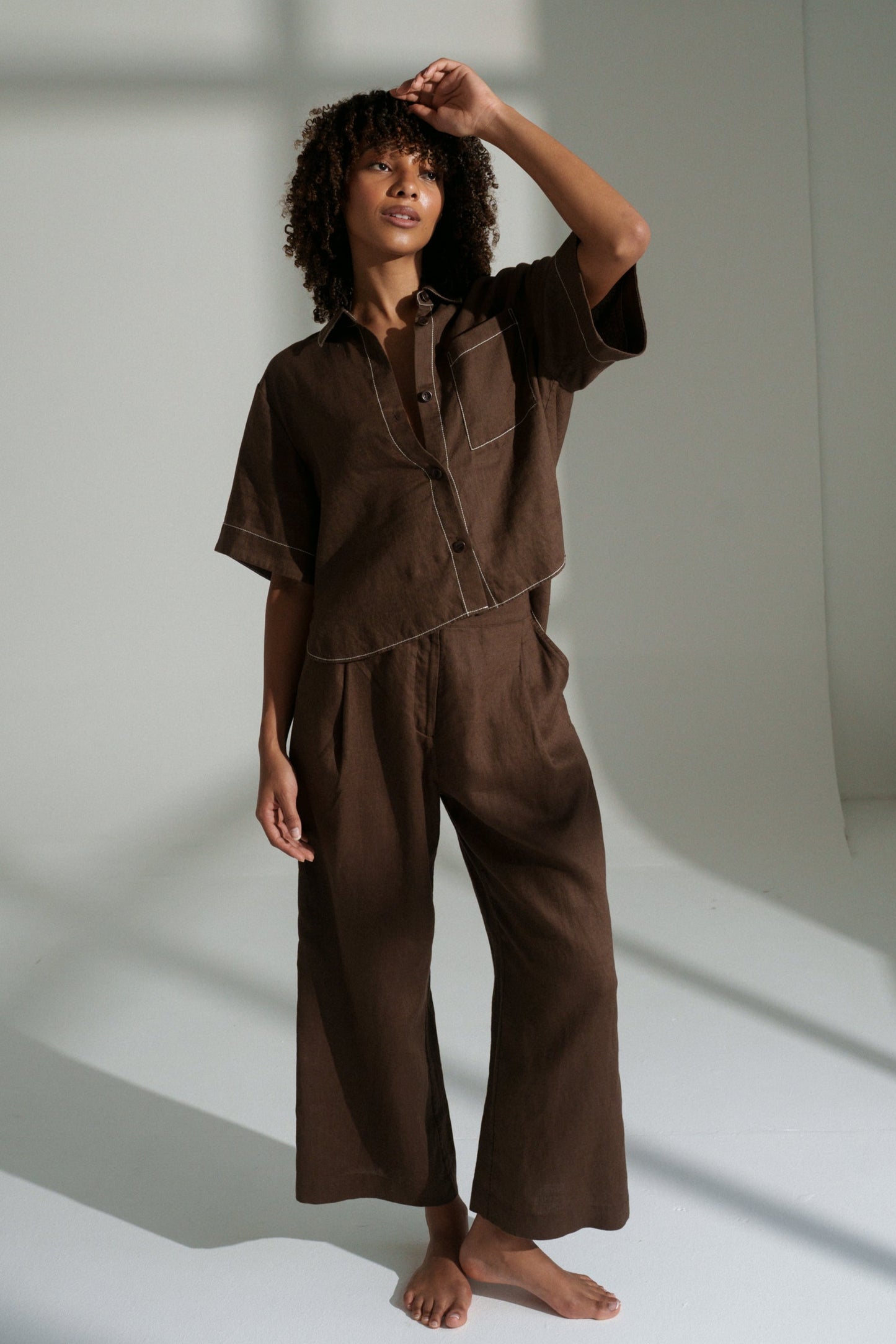LILLY PILLY woman wearing Rika pants and Nika shirt in chocolate brown standing in a well-lit room with shadows on the wall.