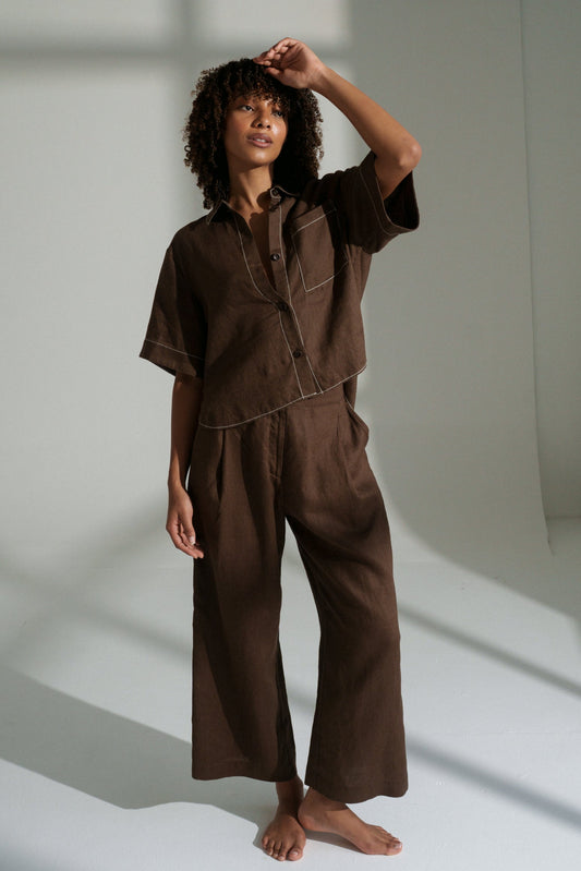 LILLY PILLY woman wearing Rika pants and Nika shirt in chocolate brown standing in a well-lit room with shadows on the wall.